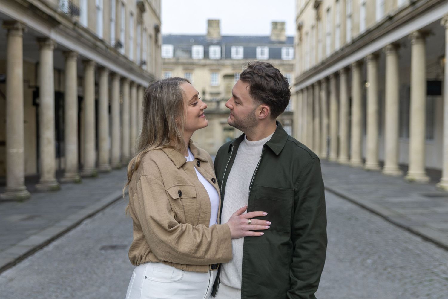 Couple sharing a romantic moment in a historic colonnade courtyard, woman in beige jacket, man in green shirt.