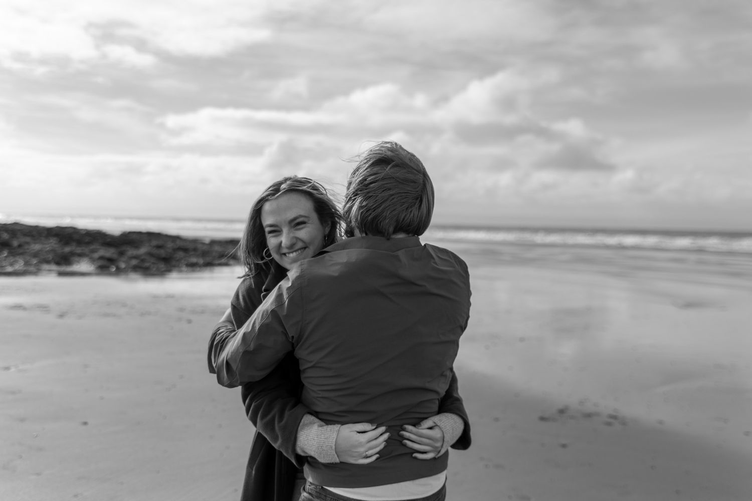 Two people embrace on a windswept beach in black and white.