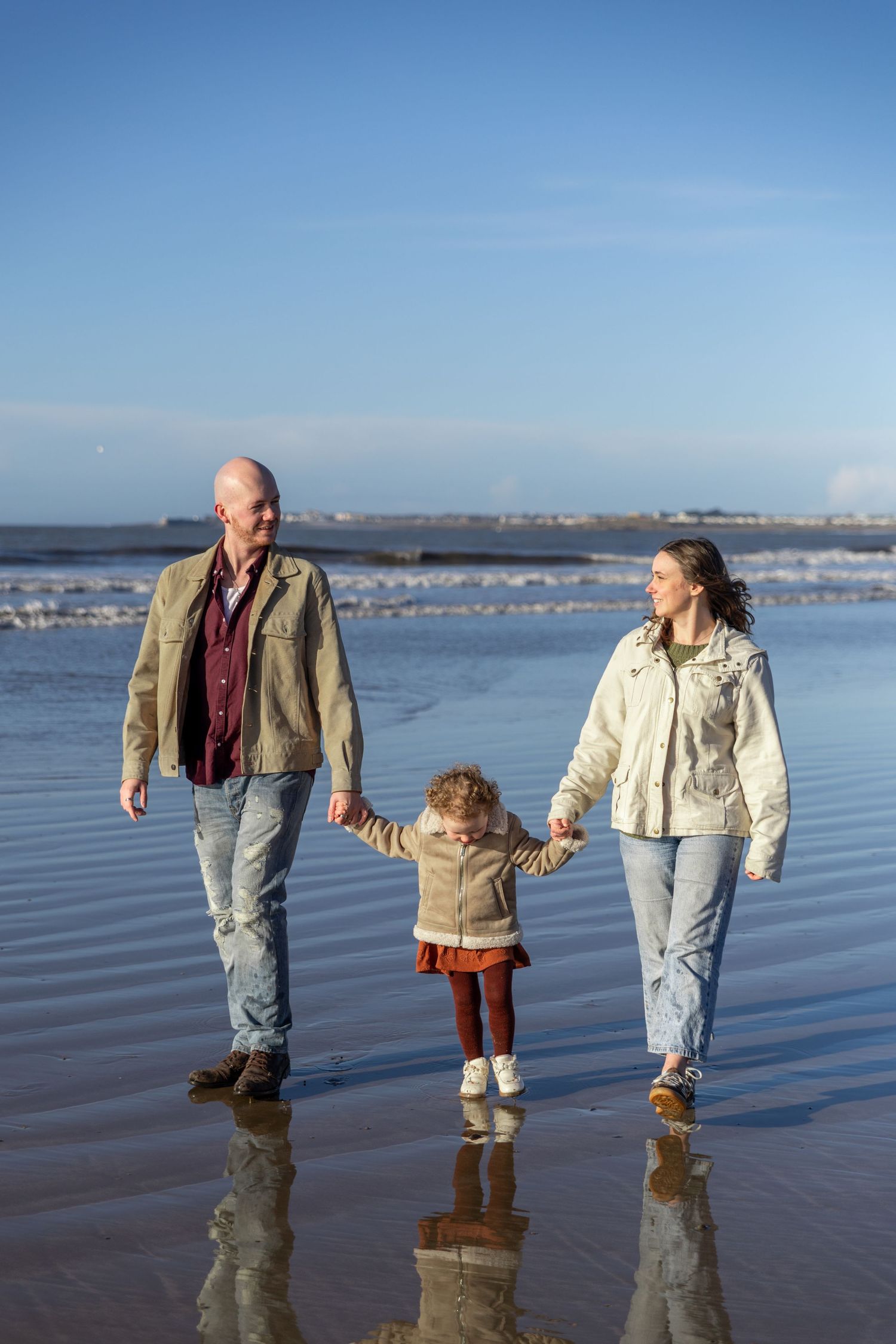 Three people walking through shallow ocean water at low tide with a coastal town visible in the background.