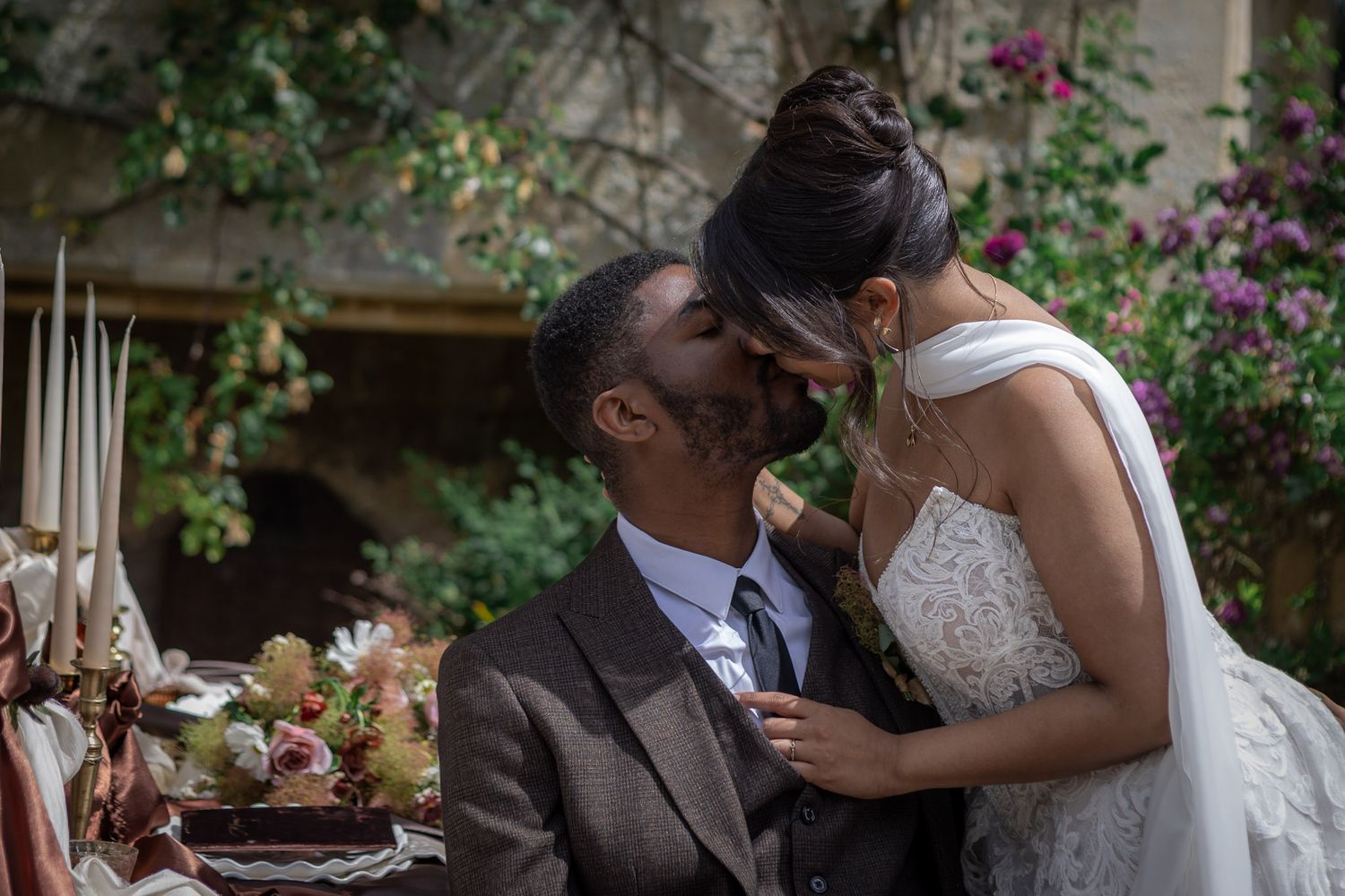 A bride and groom share a romantic kiss at their outdoor wedding reception, surrounded by lush greenery and floral arrangements.