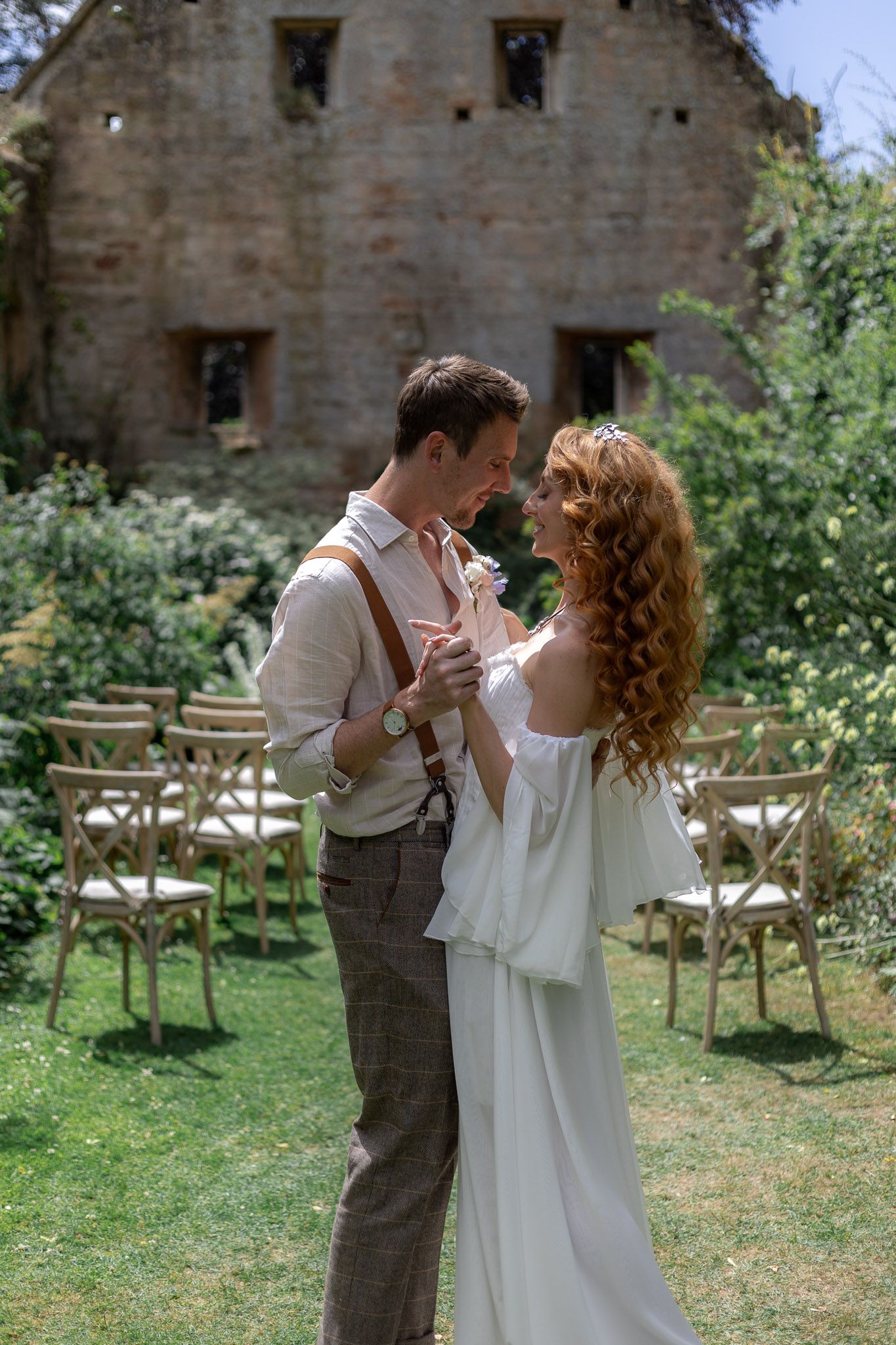A couple shares a romantic moment on a lawn with wooden chairs and stone architecture in the background.