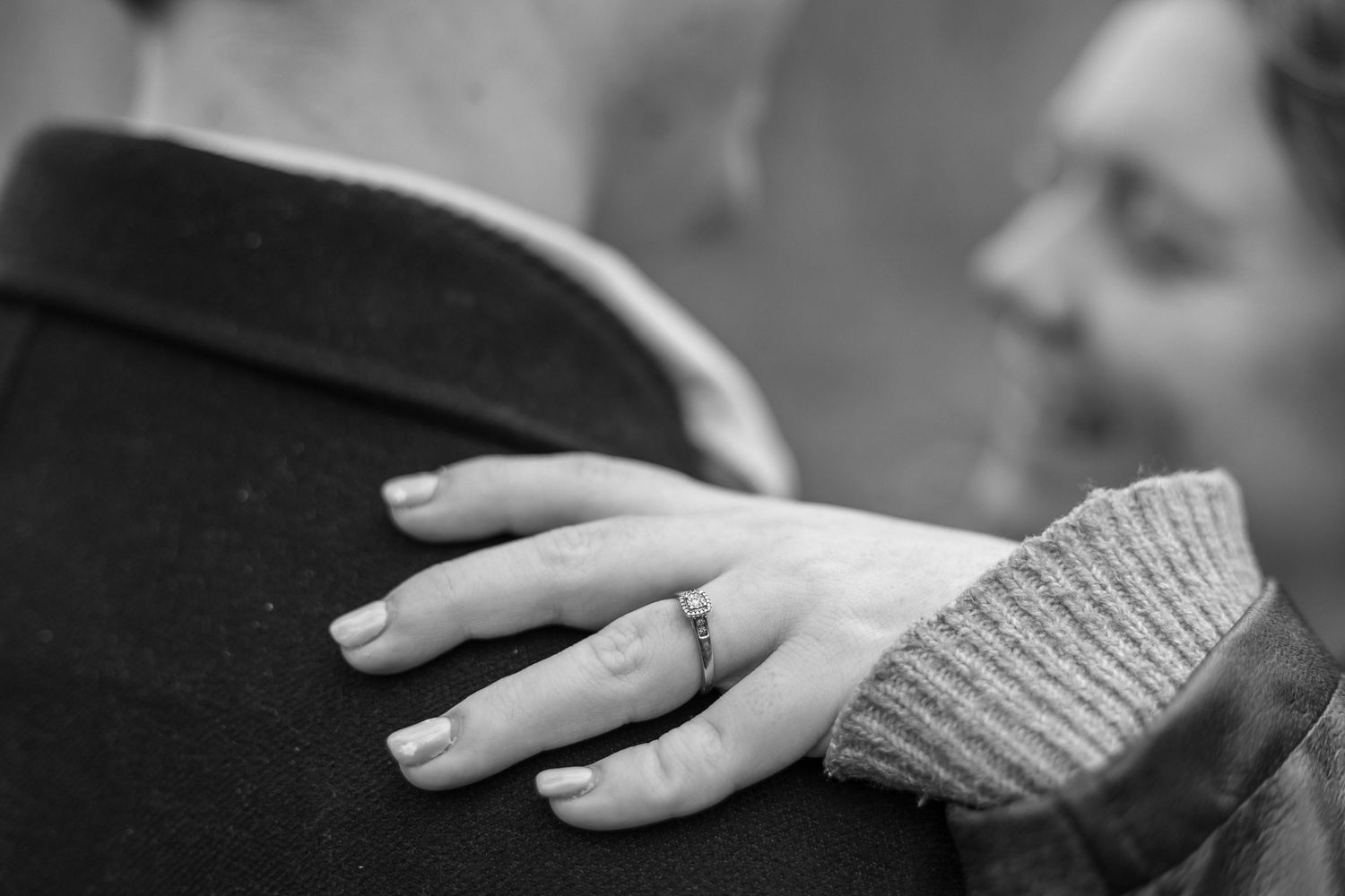 Black and white photo of a woman's hand with an engagement ring resting on a man's shoulder.