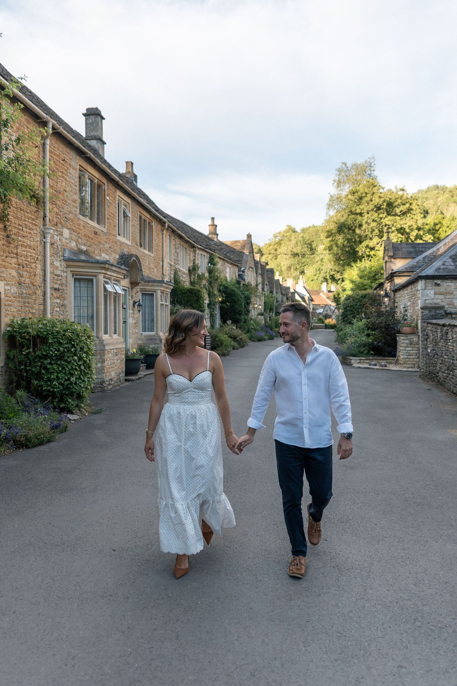 A couple holds hands walking down a charming Cotswolds village lane, woman in white dress, man in white shirt.
