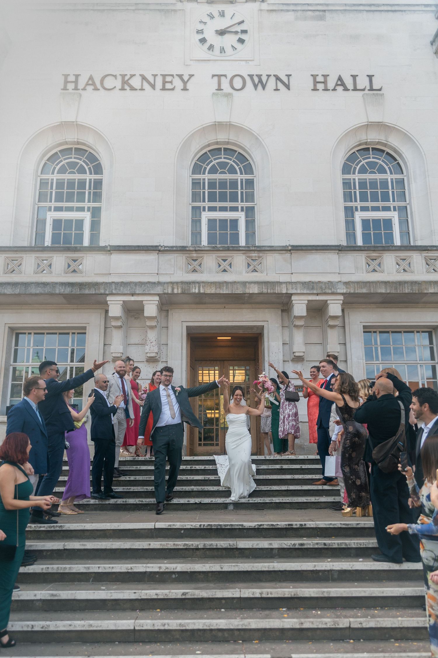 Newlyweds exit Hackney Town Hall on wedding steps, celebrated by guests throwing confetti in London.