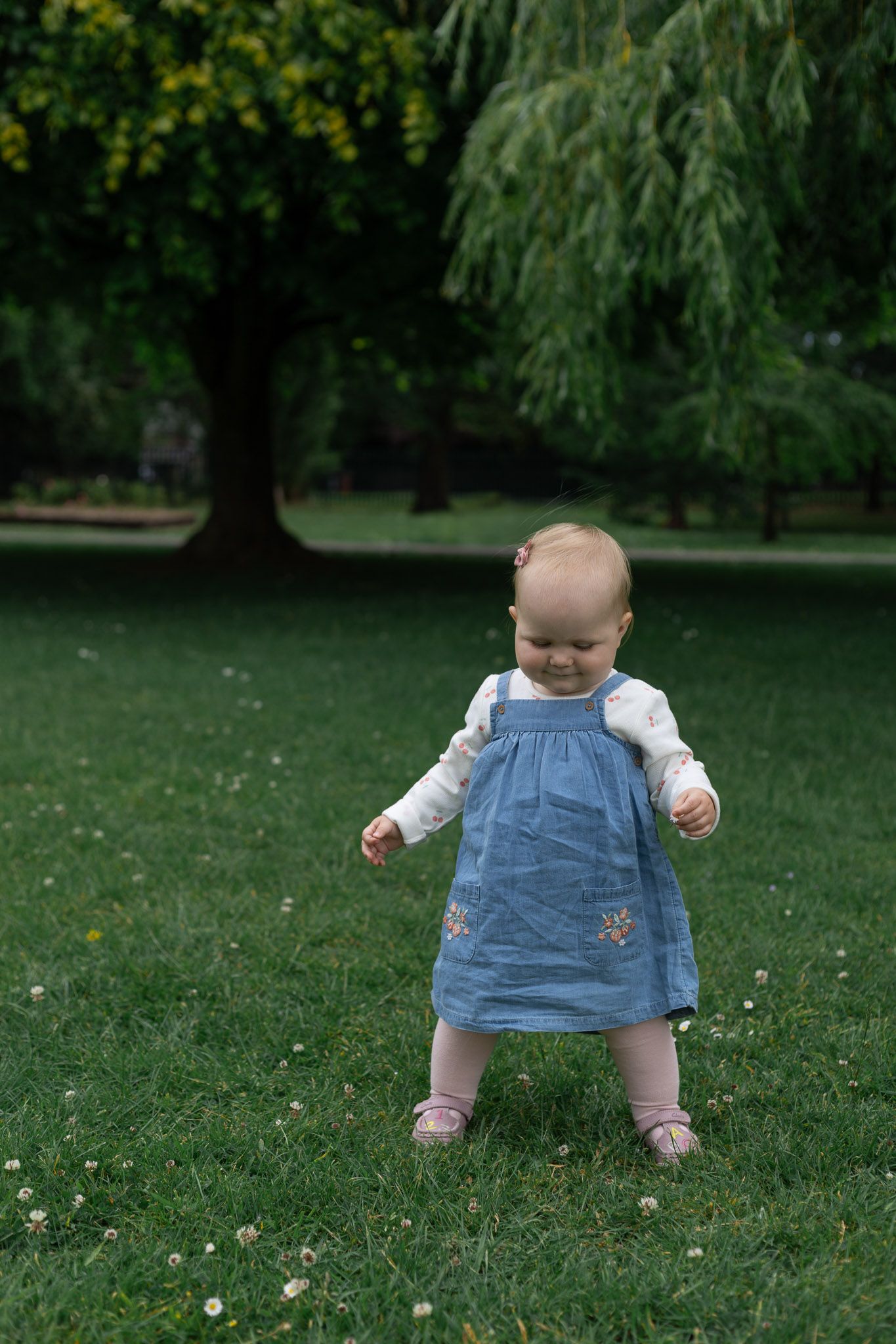 Small child in denim dress playing on grass with white flowers.