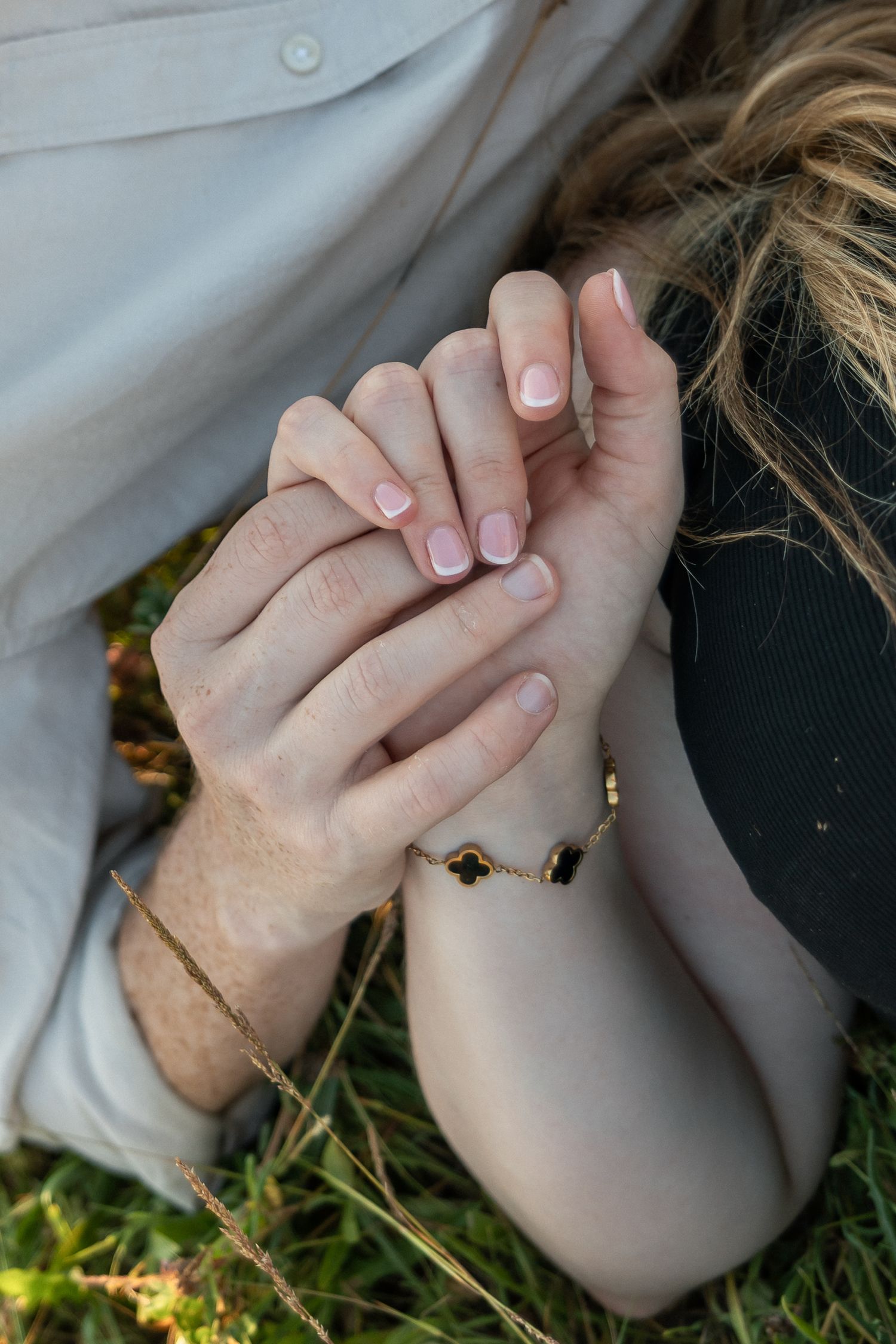 Close-up of two people holding hands on grass, one wearing a delicate gold bracelet and French manicured nails.