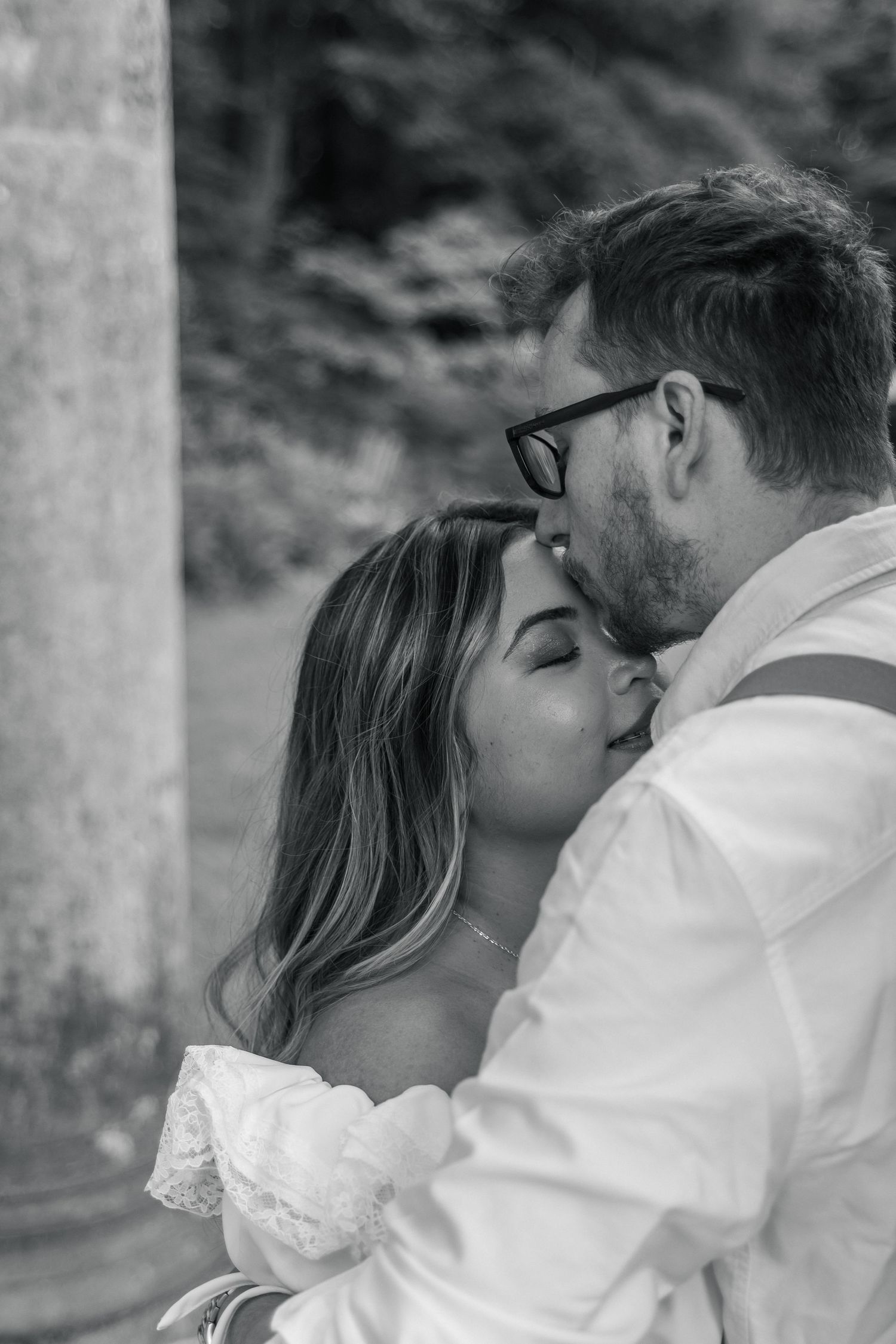 Black and white photo of a couple sharing a tender kiss, bride in lace dress and groom in white shirt with glasses.