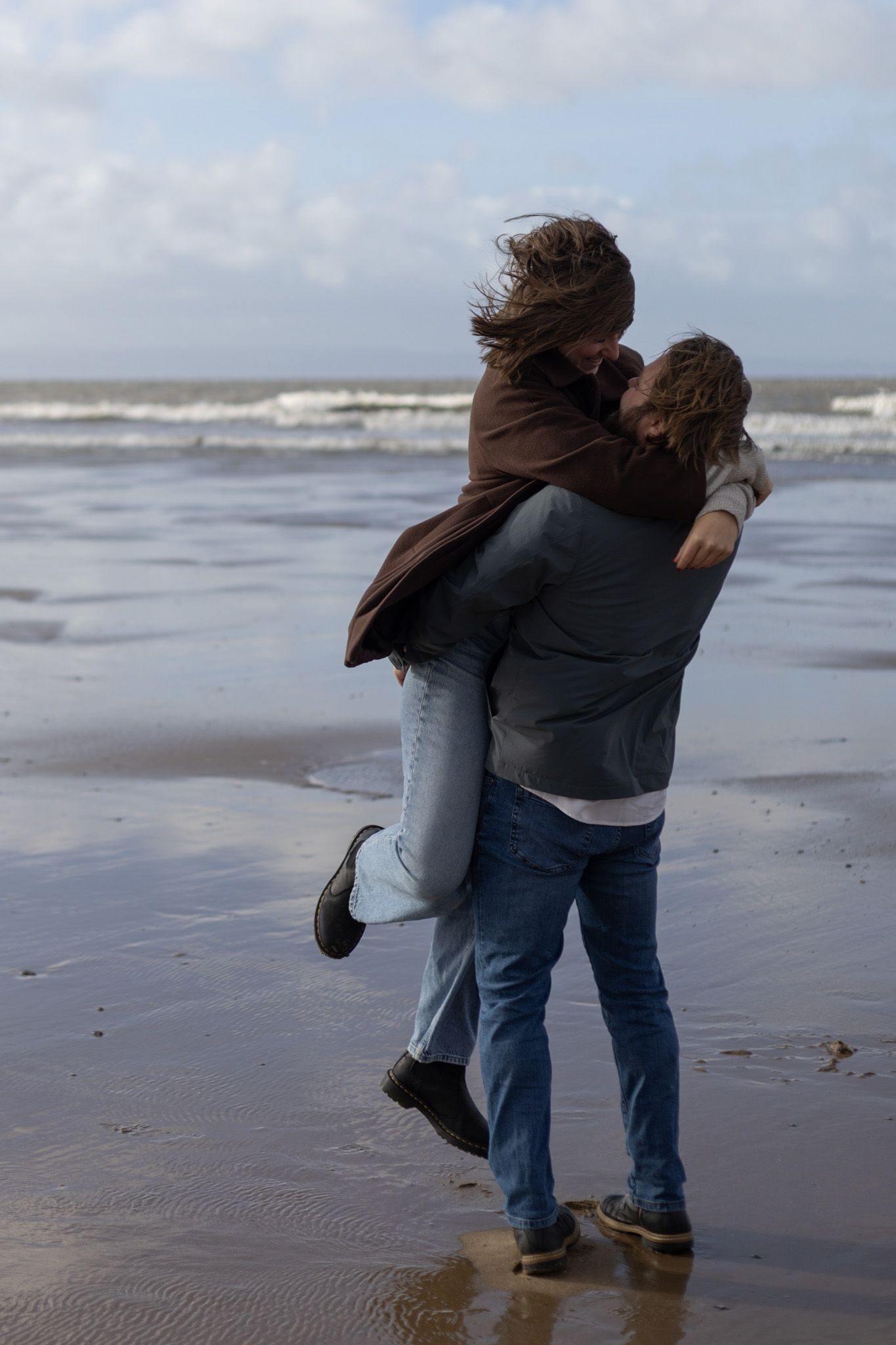 Pair walking closely together on a moody beach with waves crashing in the distance.