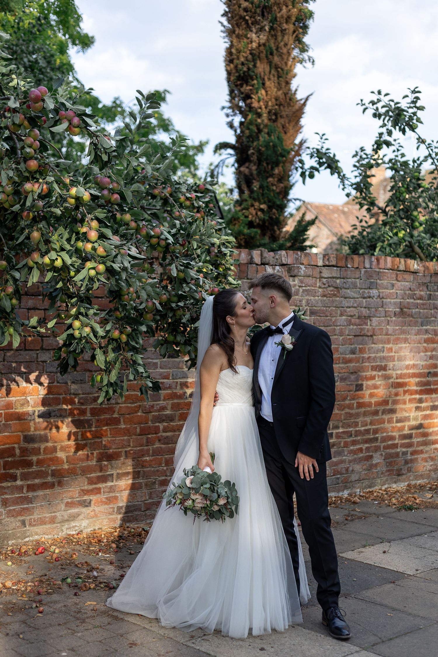 Newlywed couple shares romantic moment in garden setting with brick wall and lush greenery.