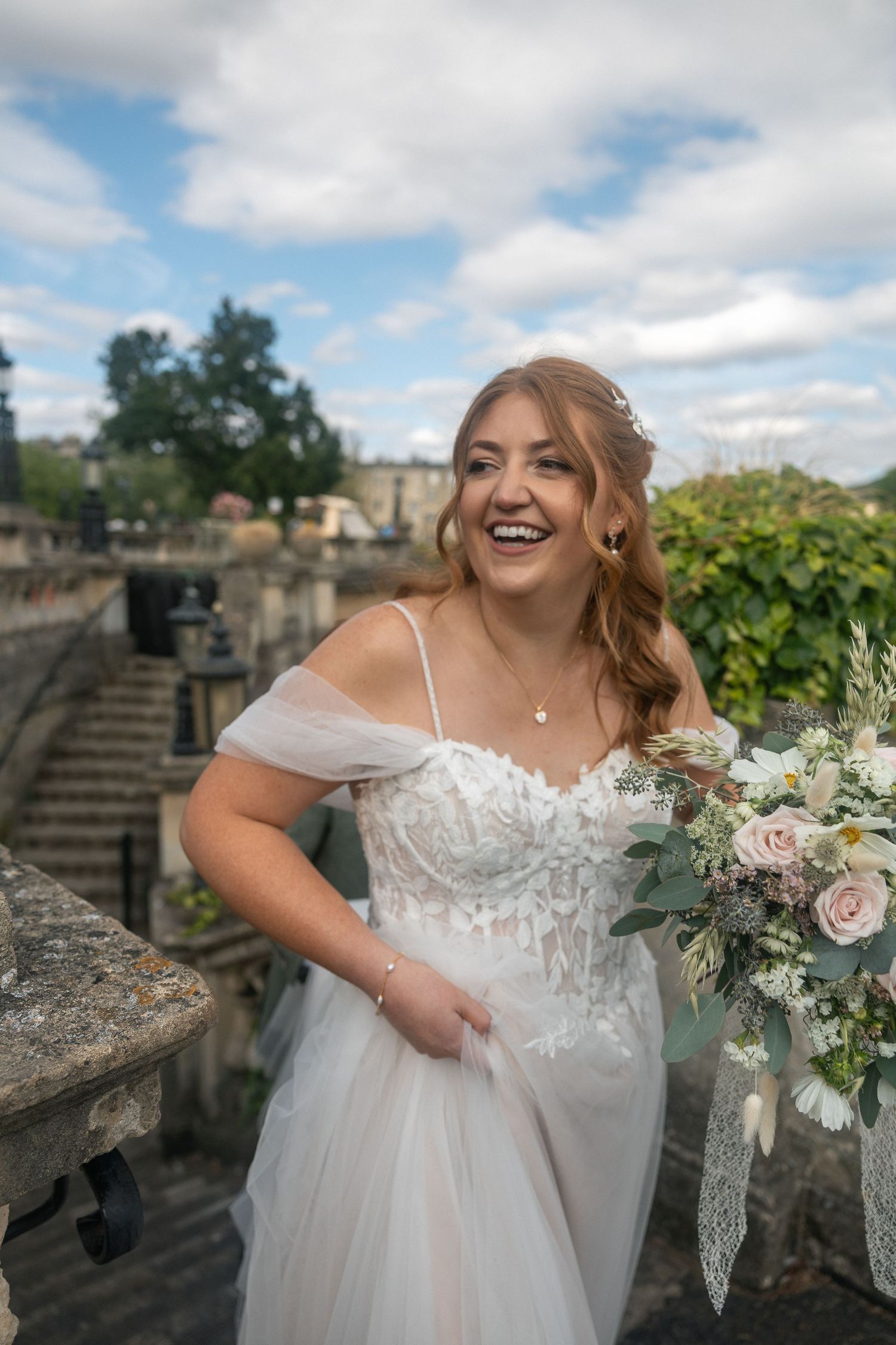 Laughing bride in off-shoulder lace wedding gown holding floral bouquet on stone balcony of Parade Gardens, Bath.