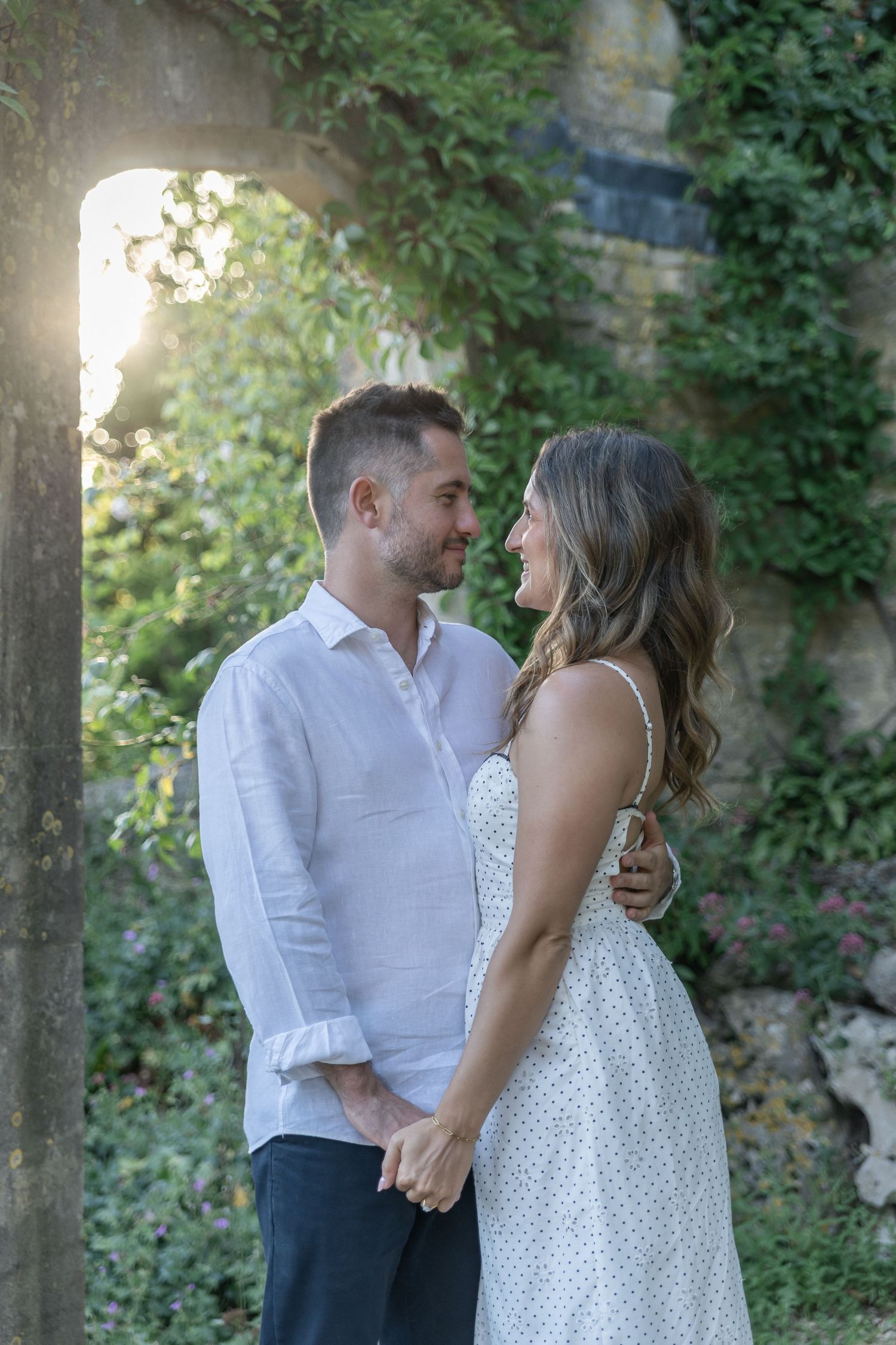 Couple holding hands and gazing at each other outdoors near stone arch with lush green ivy backdrop at golden hour.