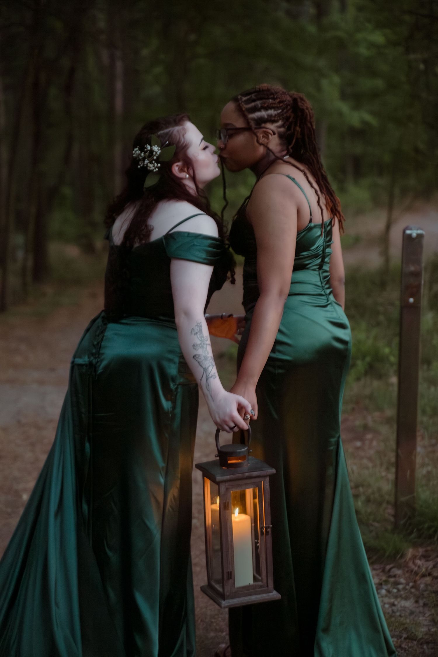 Two brides in emerald green satin gowns sharing a kiss while holding a glowing lantern in forest.