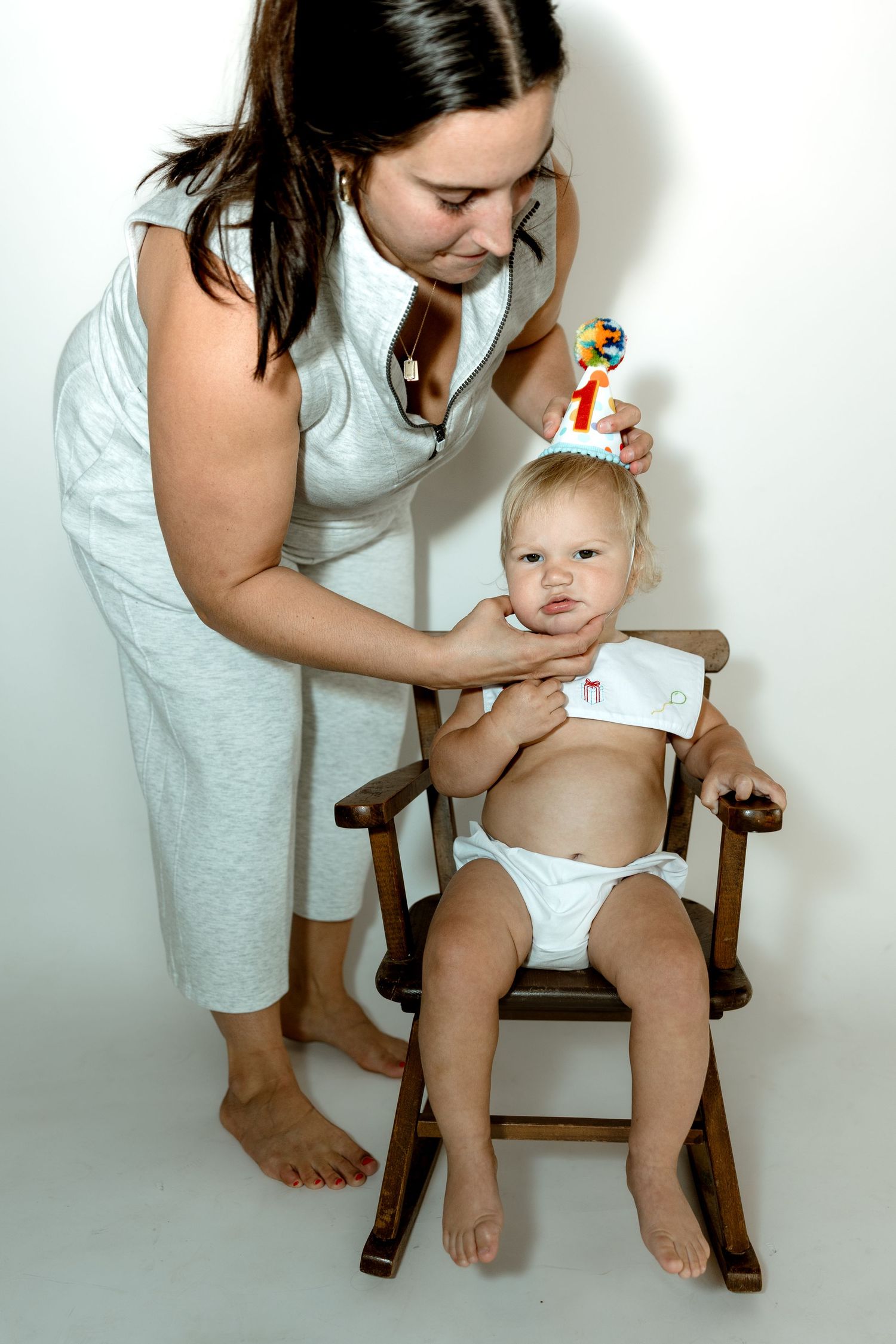 Mother placing birthday hat on baby sitting in wooden rocking chair during first birthday photo.