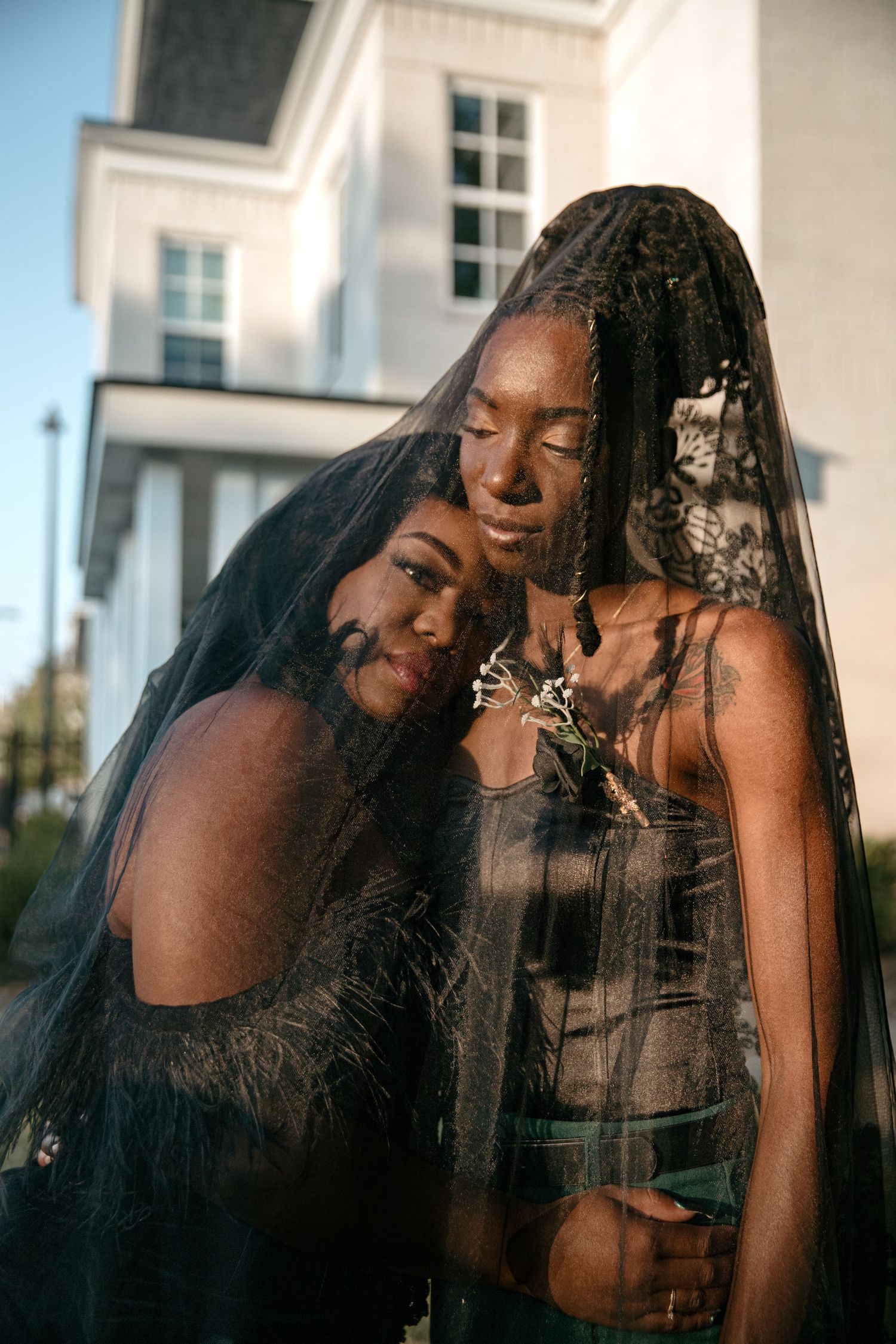 Two Black women pose together under a sheer black veil outside a white building at sunset.