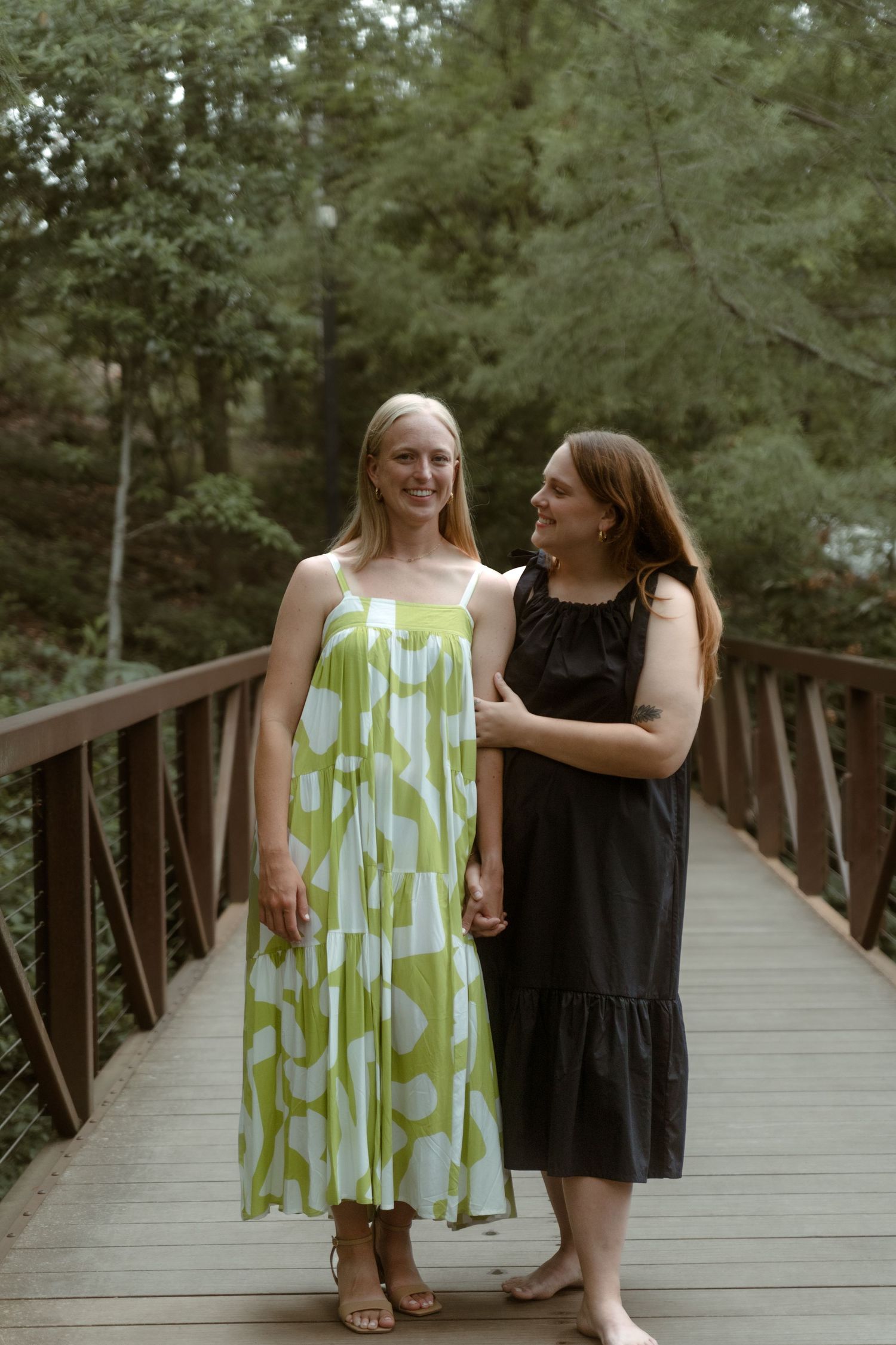 Two women in summer dresses smile together on a wooden bridge in a forest.