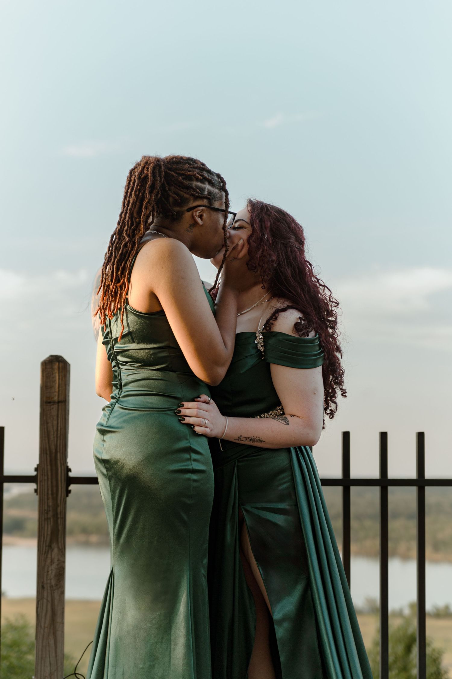 Two women in matching green satin gowns share a kiss outdoors at sunset.