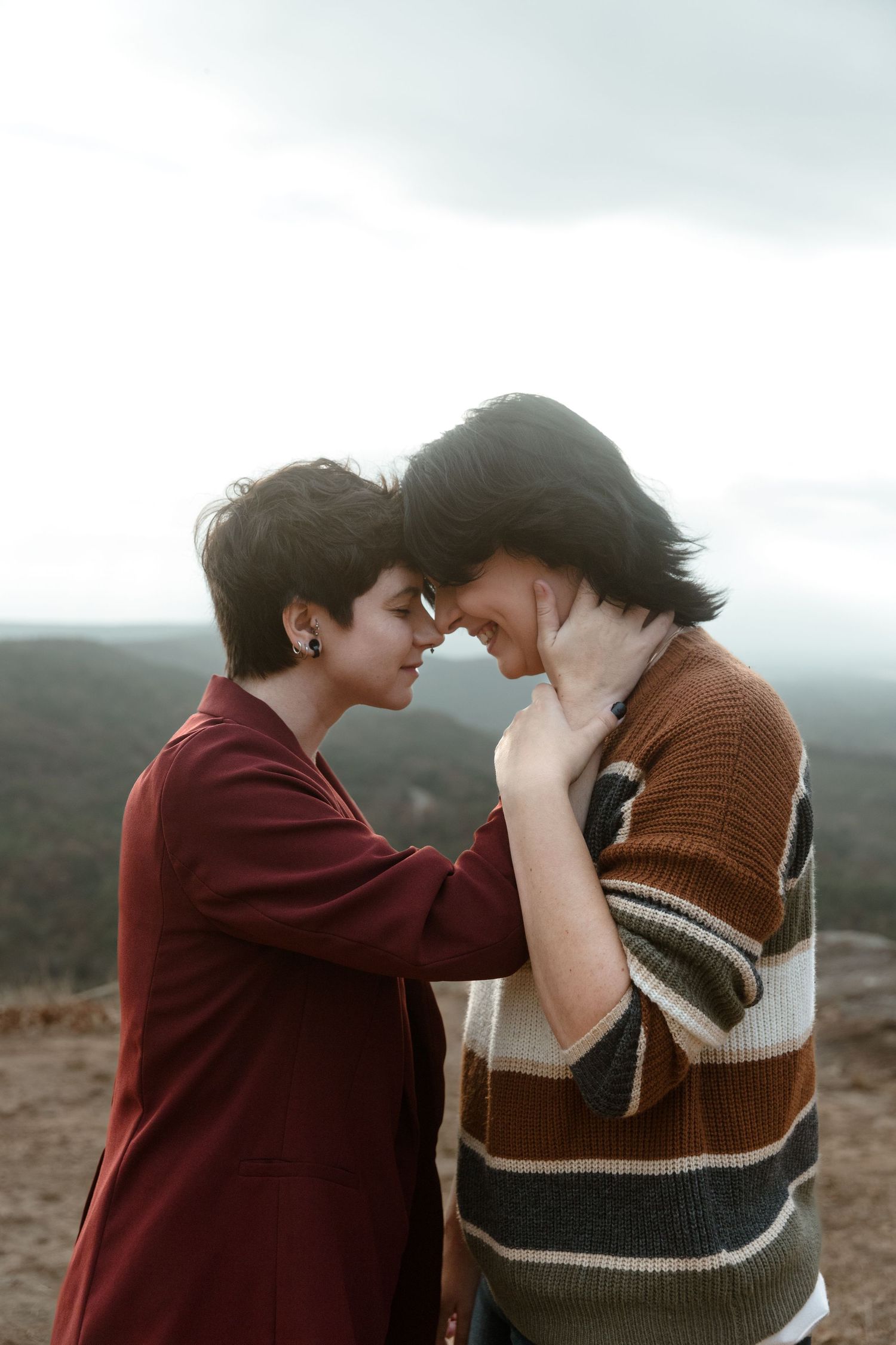Couple touching foreheads tenderly on mountain overlook under cloudy sky.