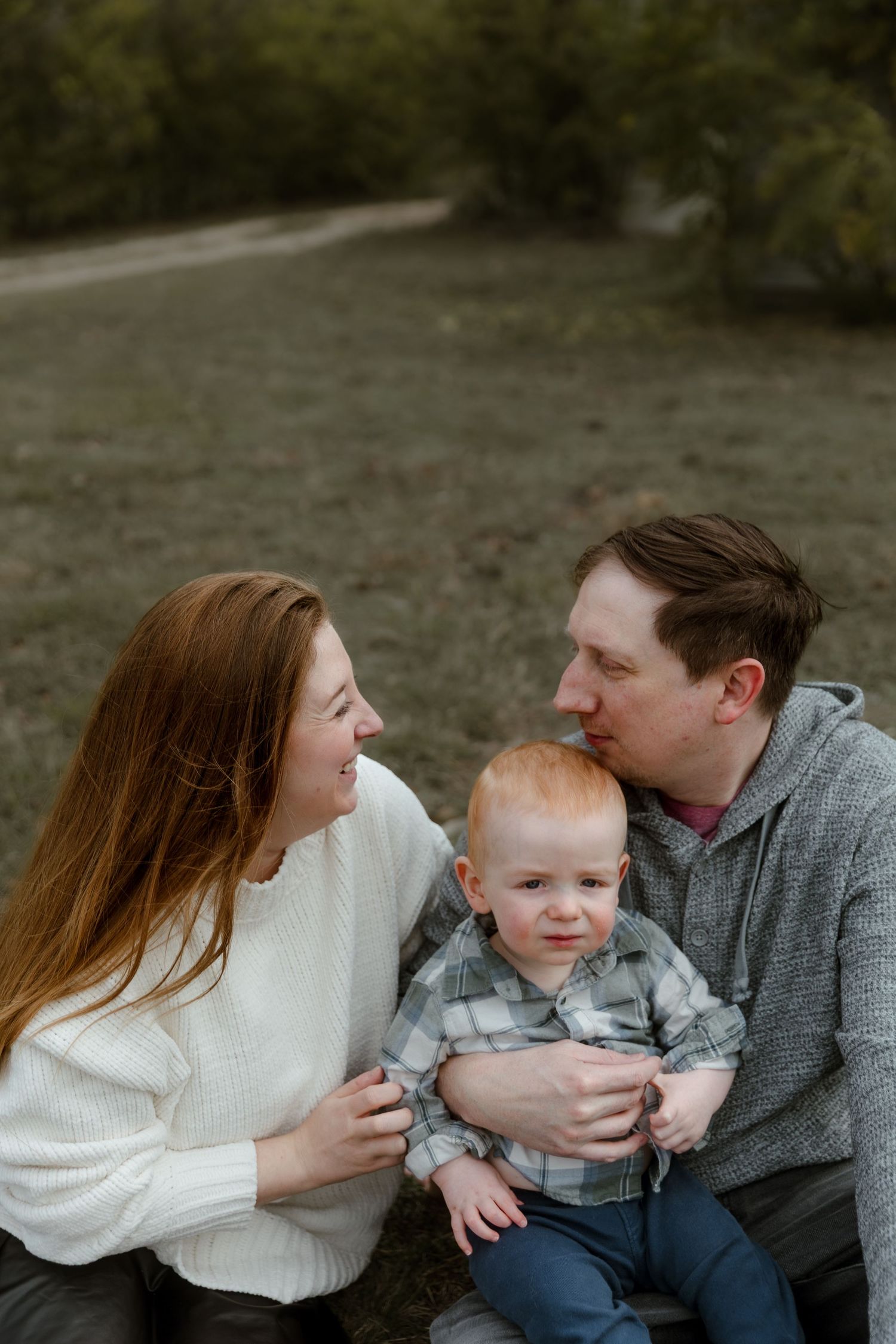 A red-haired mother and father sit outdoors gazing at each other while holding their baby.