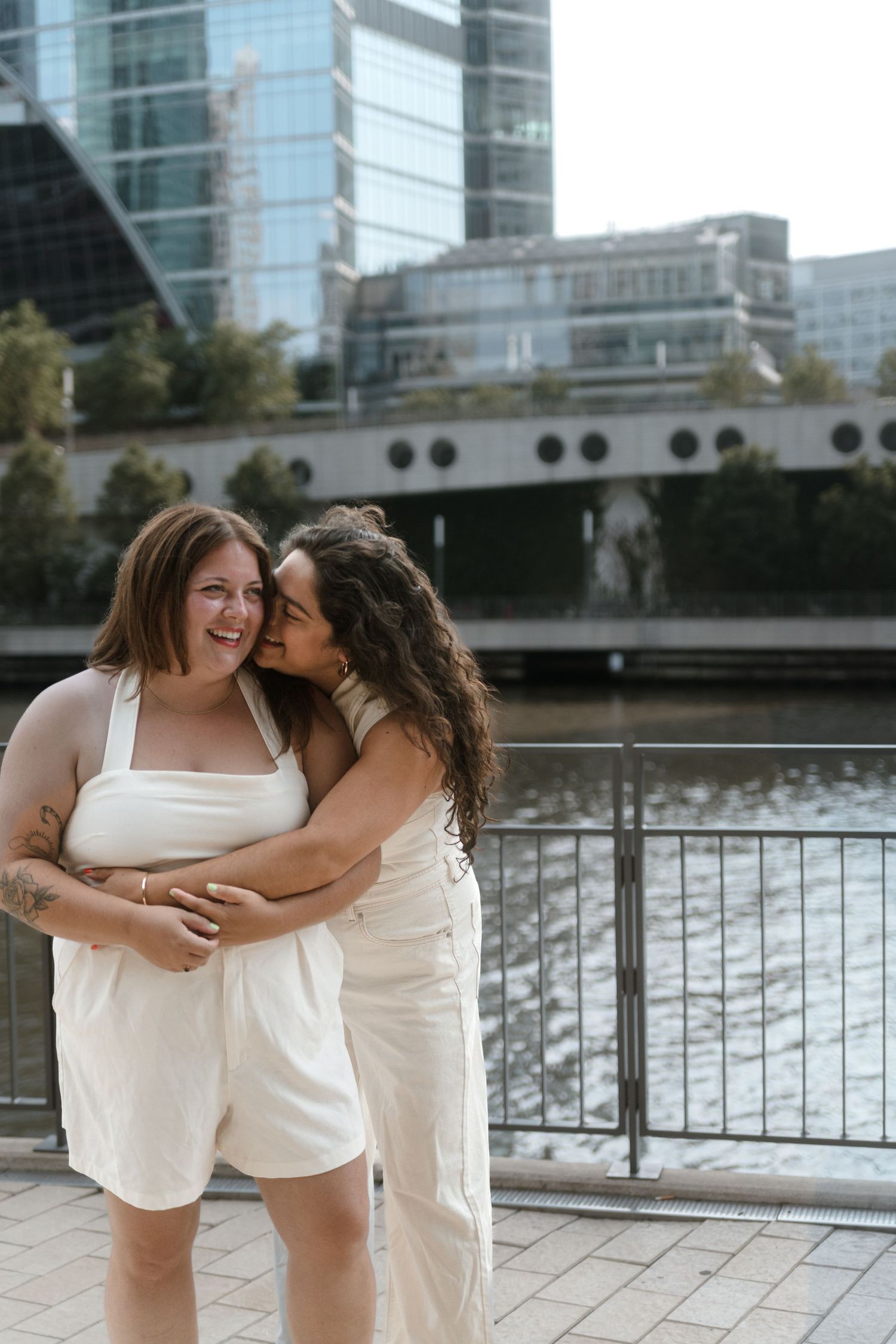 Two women in white outfits embrace and laugh together along an urban waterfront.