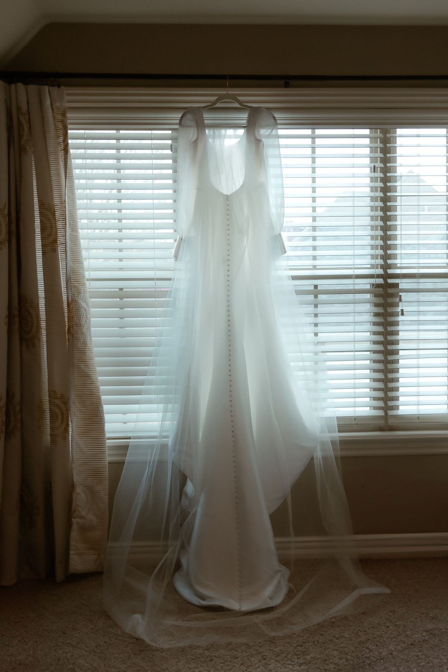 A white wedding gown with a long veil hangs elegantly in front of a sunlit window.