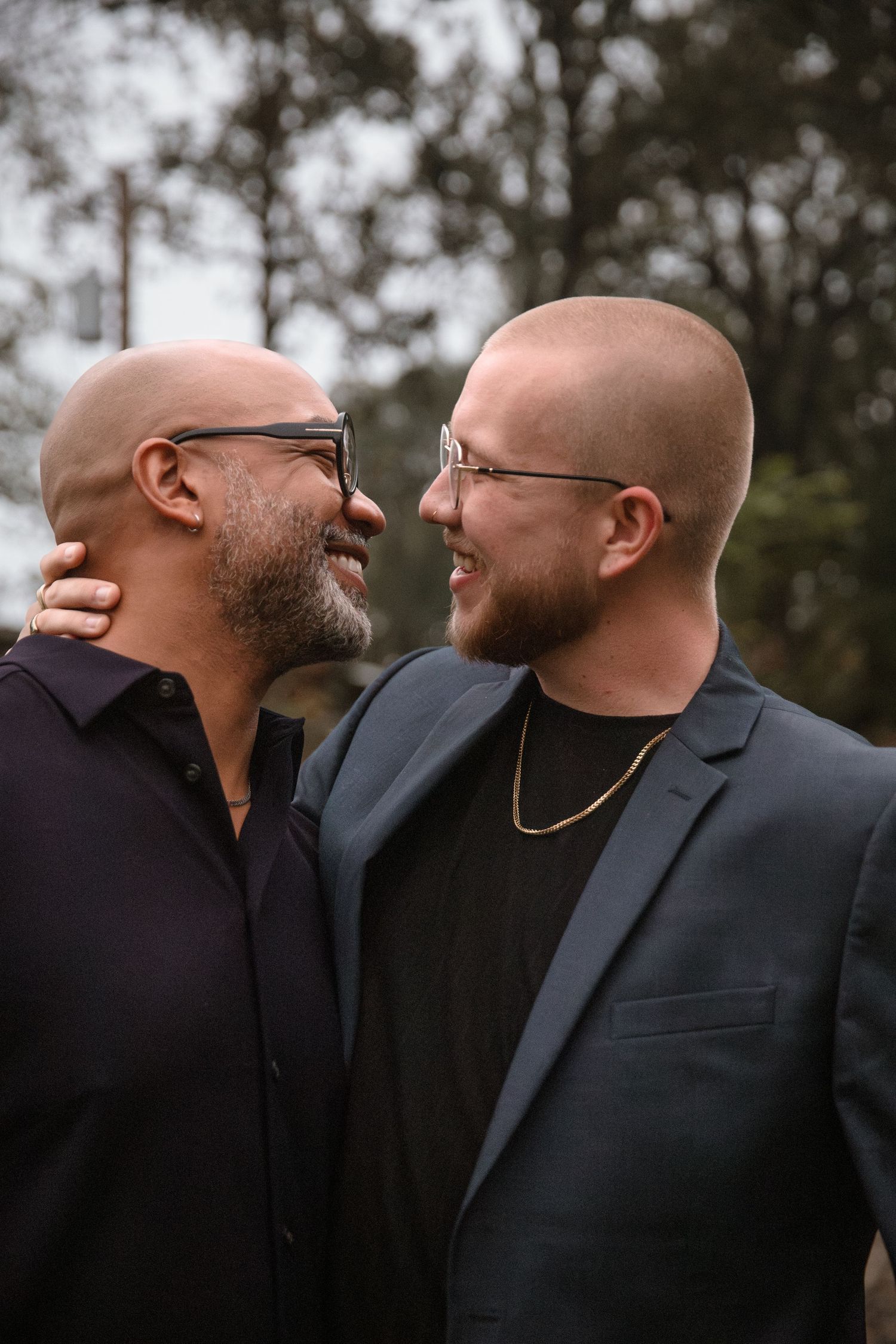 Two bald men in suits share a joyful moment, smiling at each other during a wedding celebration outdoors.