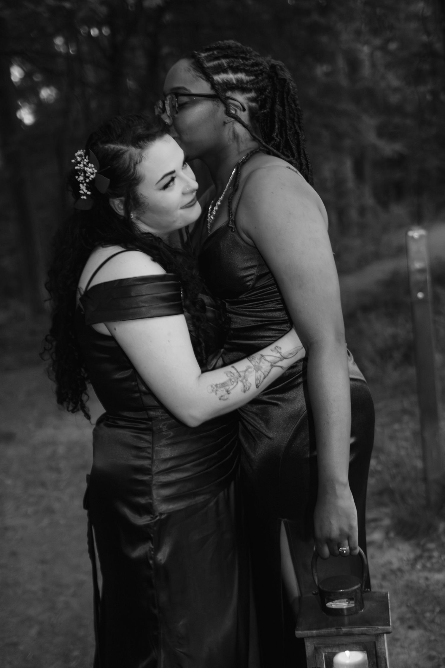 Black and white photo of two women in satin gowns embracing in a forest setting.