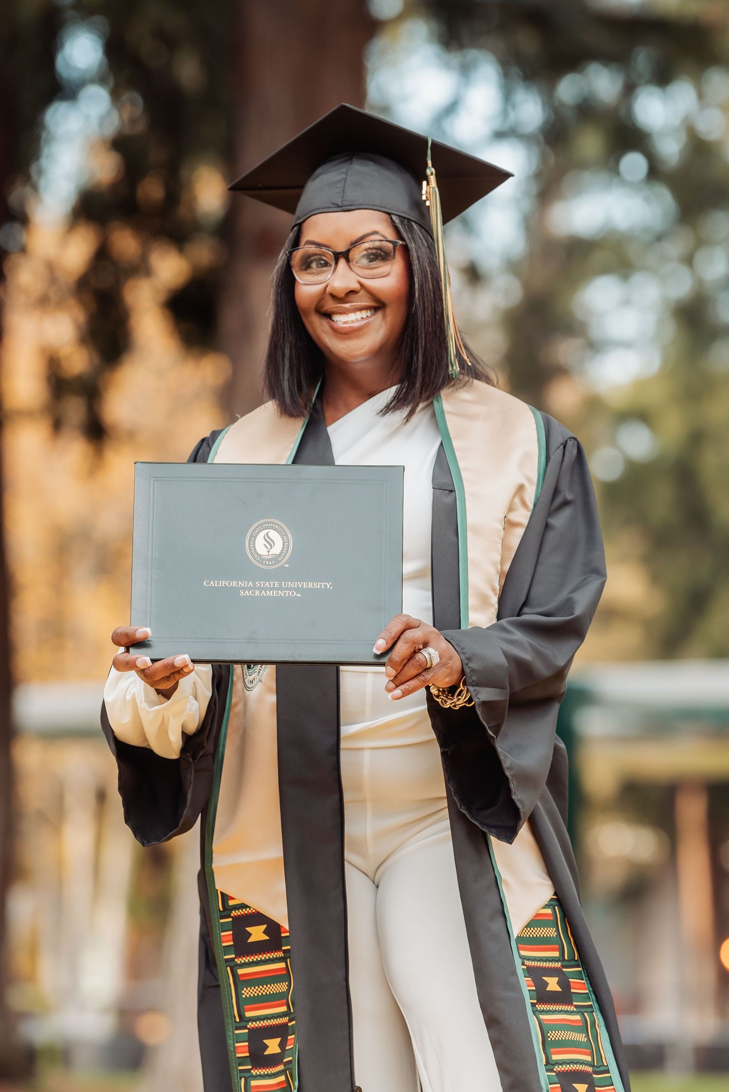 Graduate in black cap and gown with academic stole displays diploma during outdoor graduation photo session.