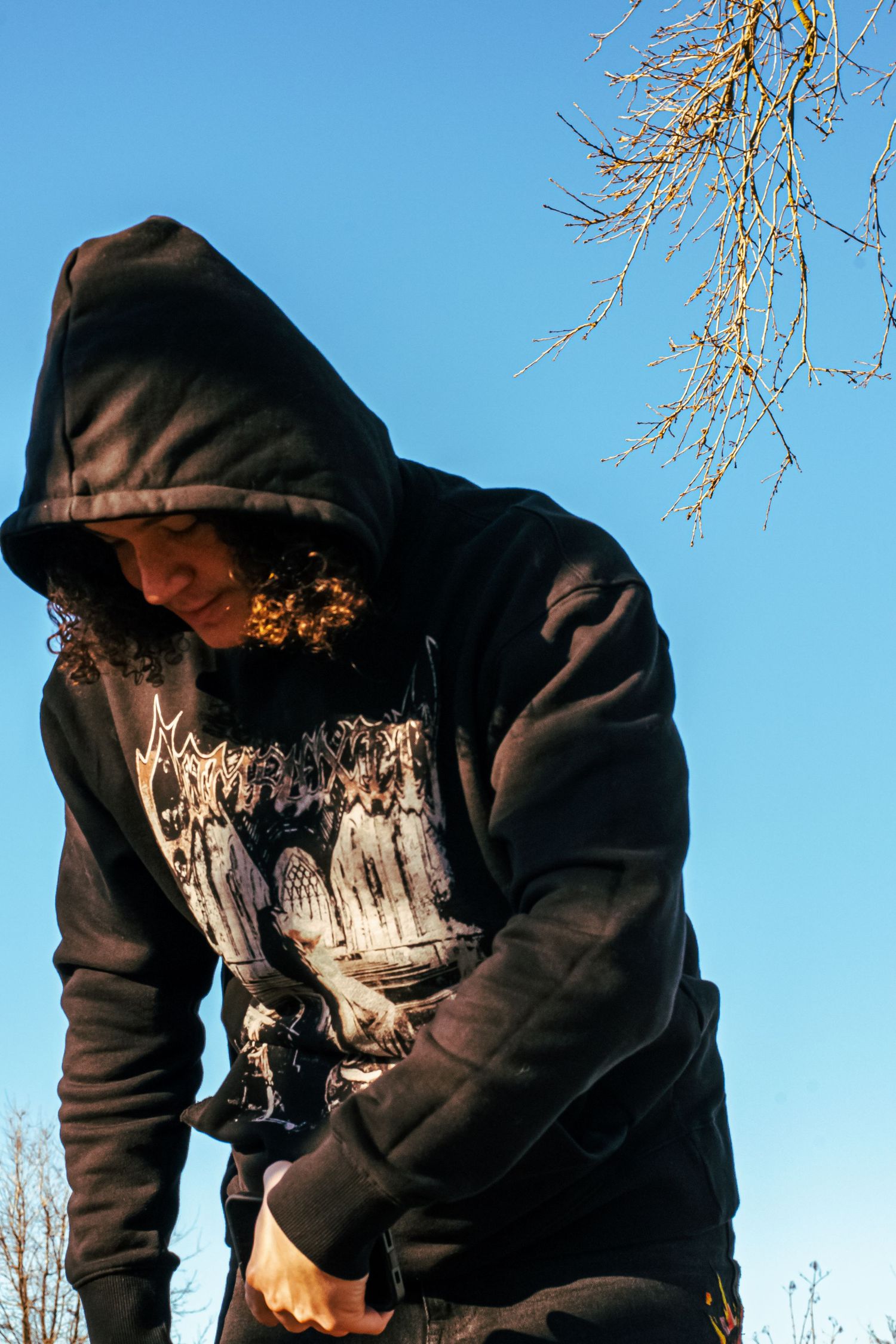 Someone wearing a black hooded sweatshirt with a graphic design stands against a bright blue sky with bare tree branches above.