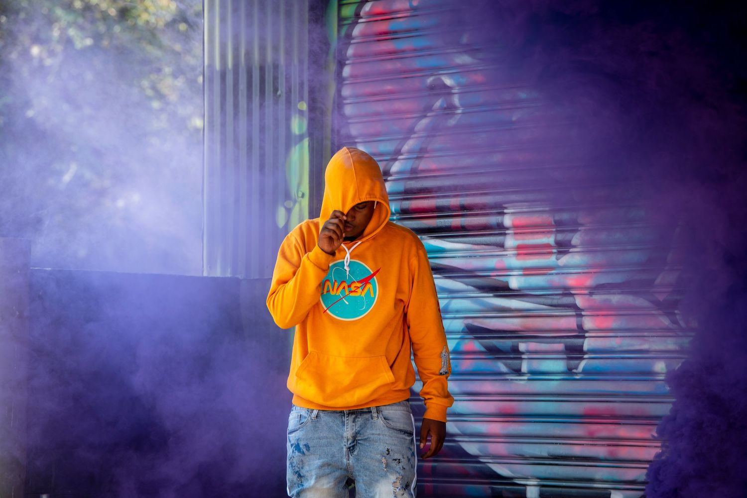Person in bright orange hoodie stands against graffitied metal door with purple lighting effects.