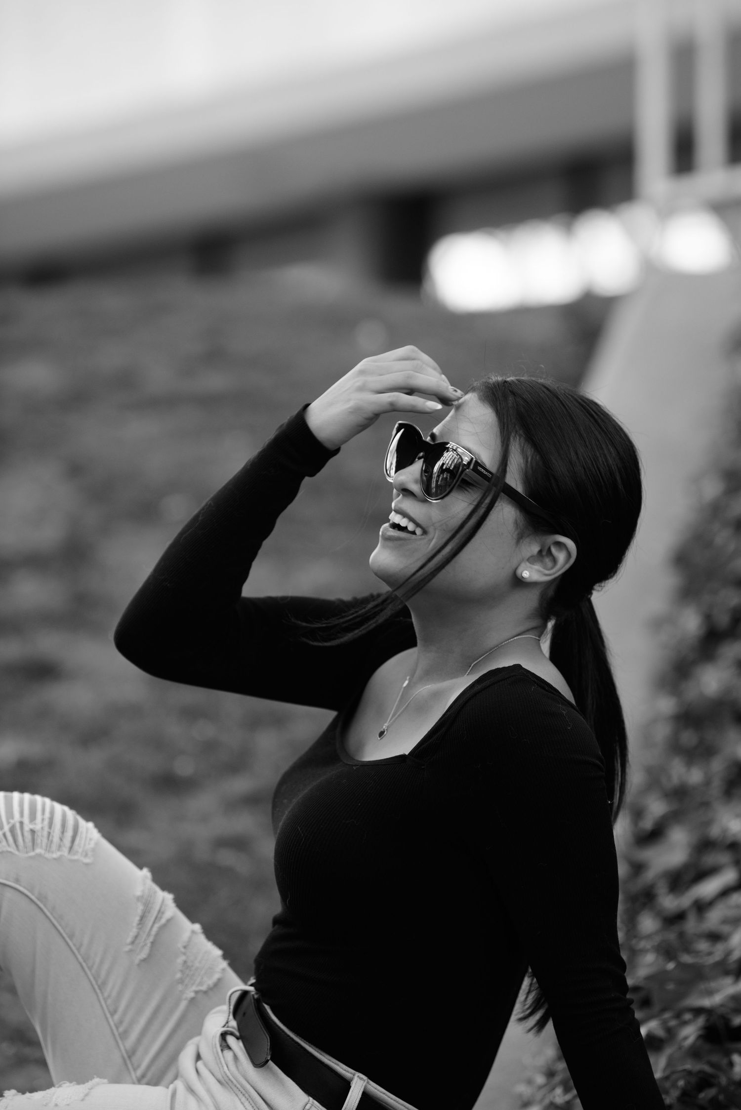 Black and white portrait of someone laughing joyfully while wearing sunglasses and a black sweater outdoors.