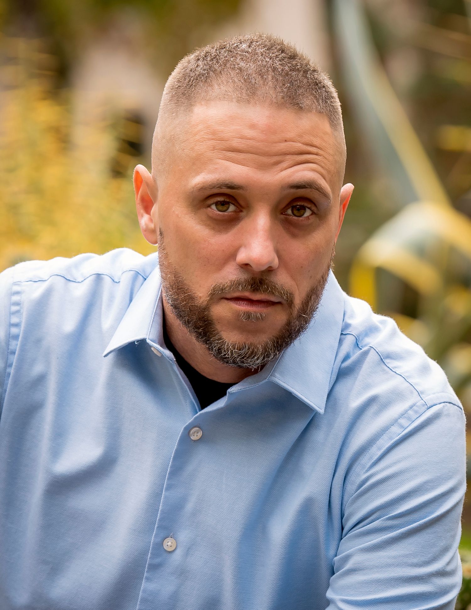 Professional headshot of person wearing light blue collared shirt against outdoor background with soft bokeh.