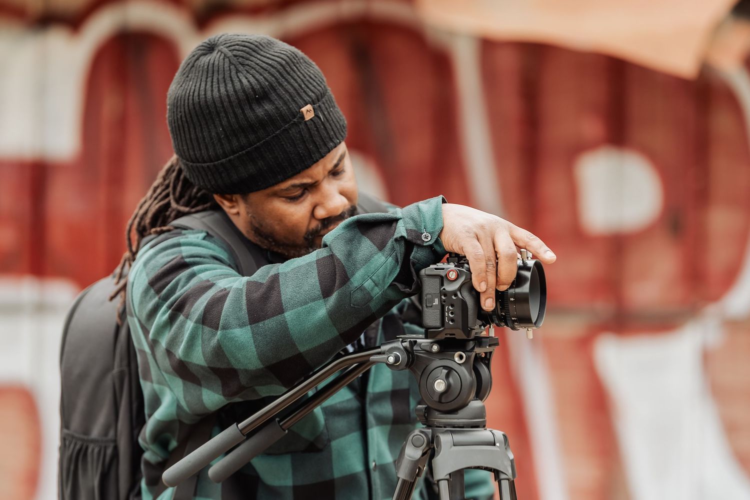 Photographer adjusting camera settings on tripod while wearing plaid jacket and beanie.
