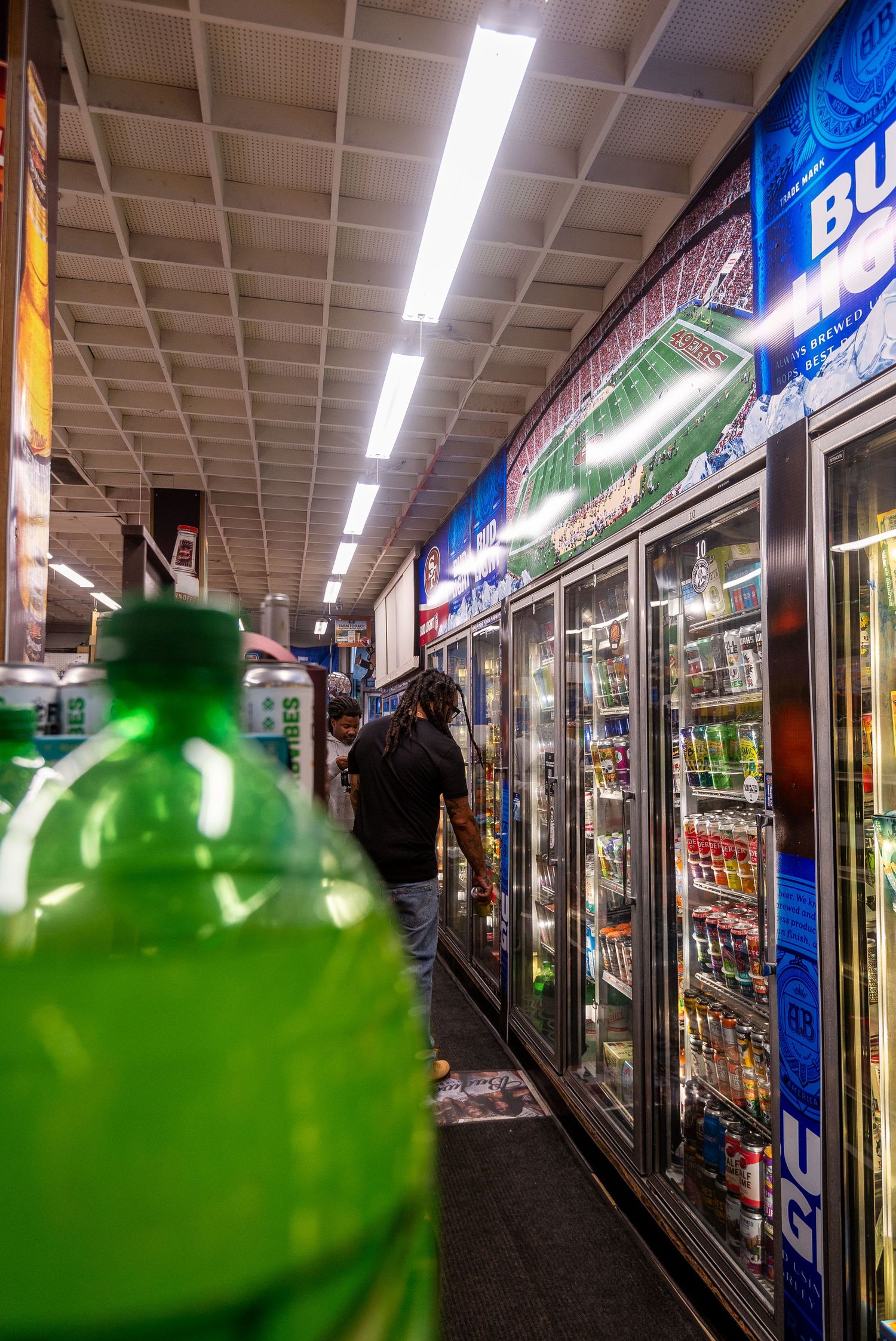 Interior view of convenience store refrigerator section with green bottle in foreground and glowing cooler displays in background.