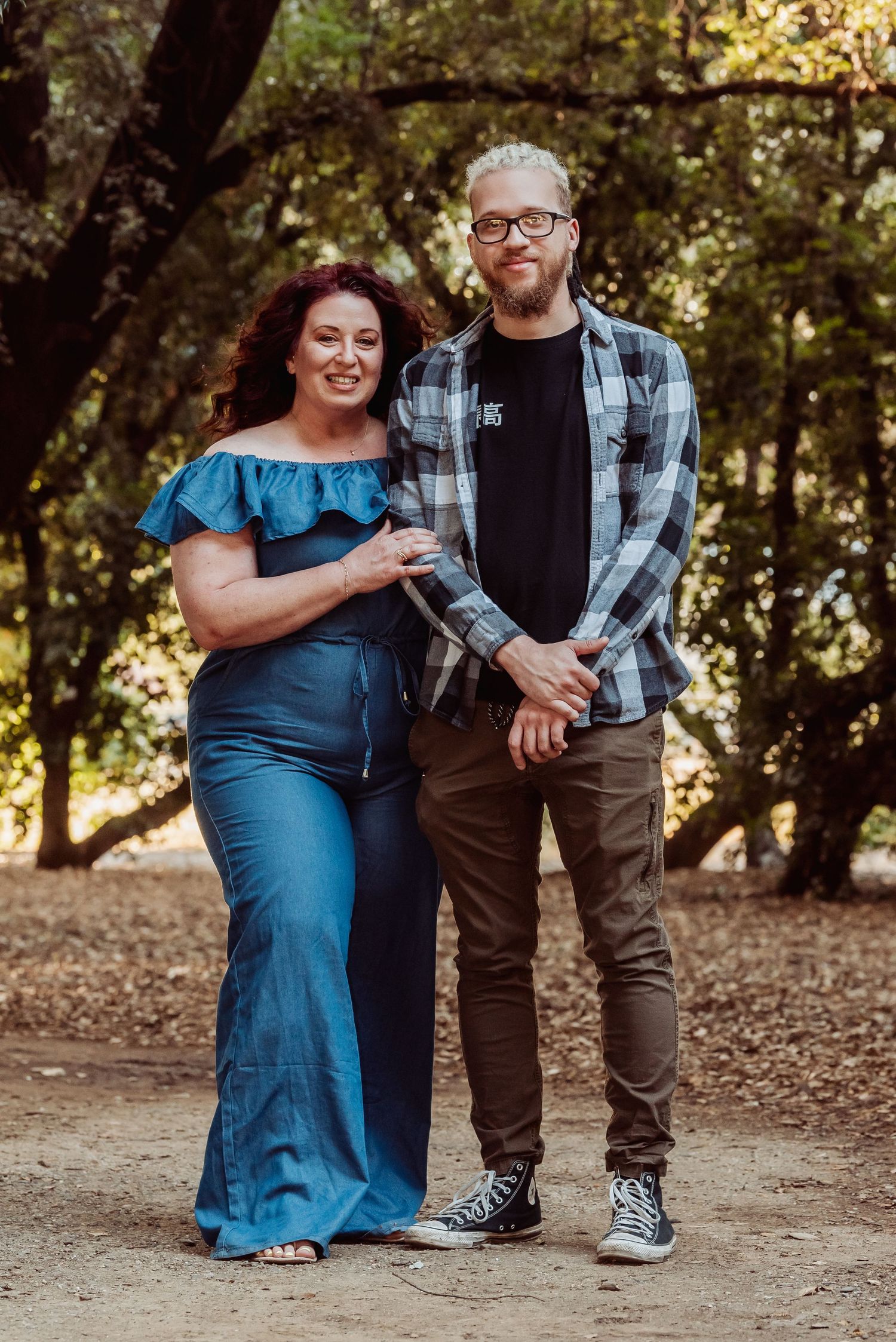 Couple poses together on woodland trail, woman in blue off-shoulder dress and man in plaid shirt and glasses.