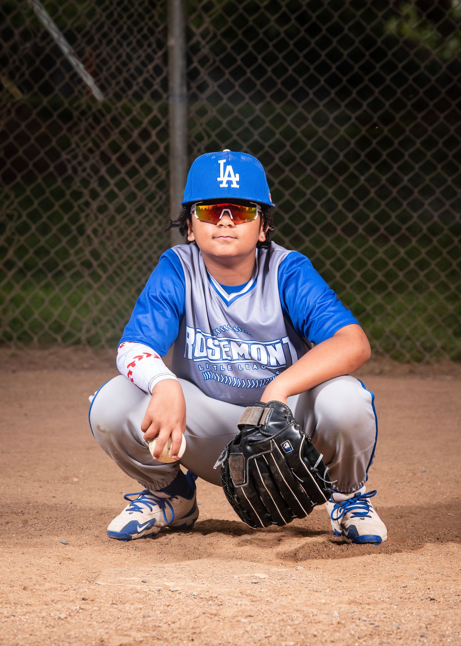 Baseball player in LA Dodgers cap and gear crouches in catching position on dirt field with glove and sunglasses.