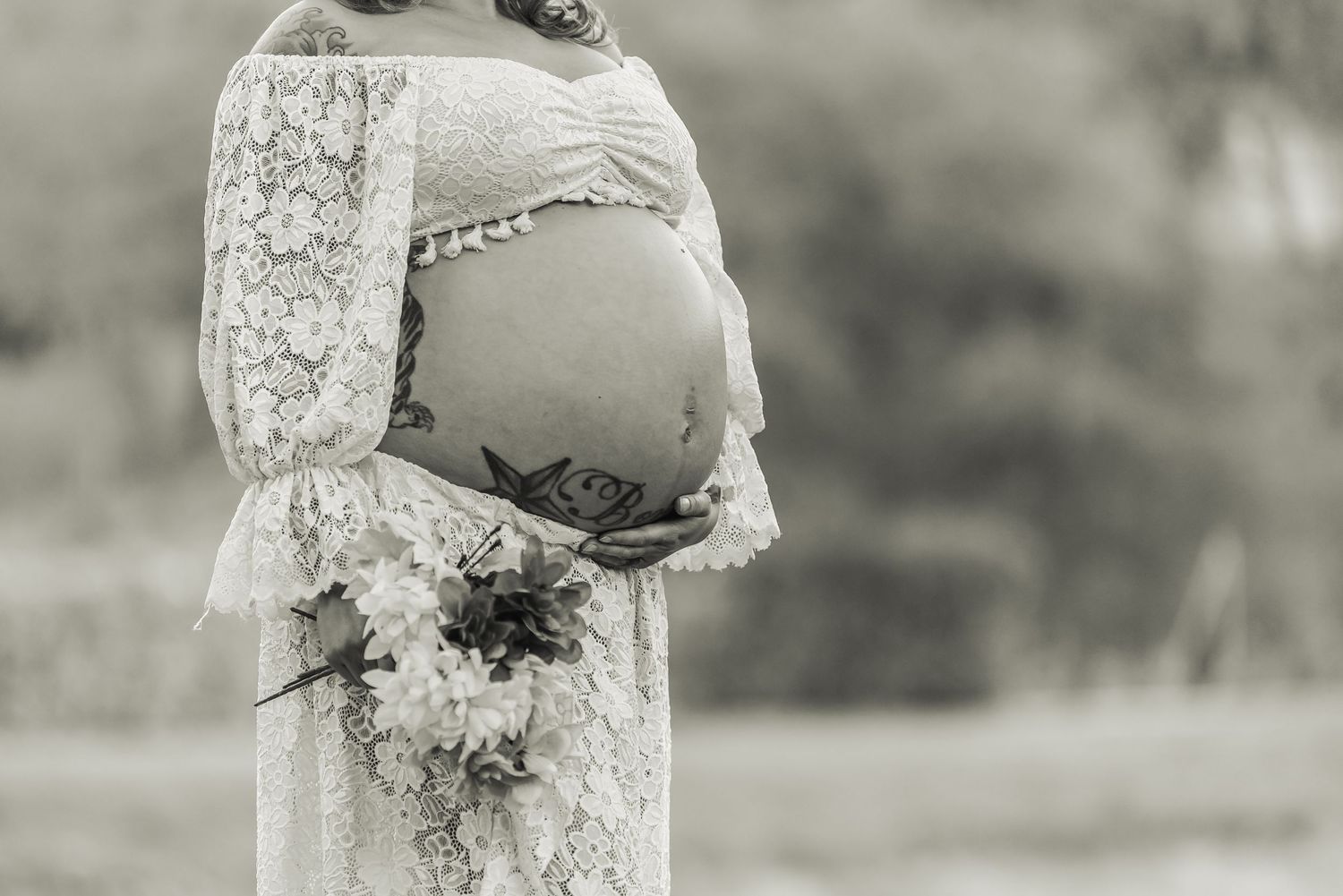 Black and white maternity photo showing a pregnant belly in lace dress while holding a floral bouquet outdoors.