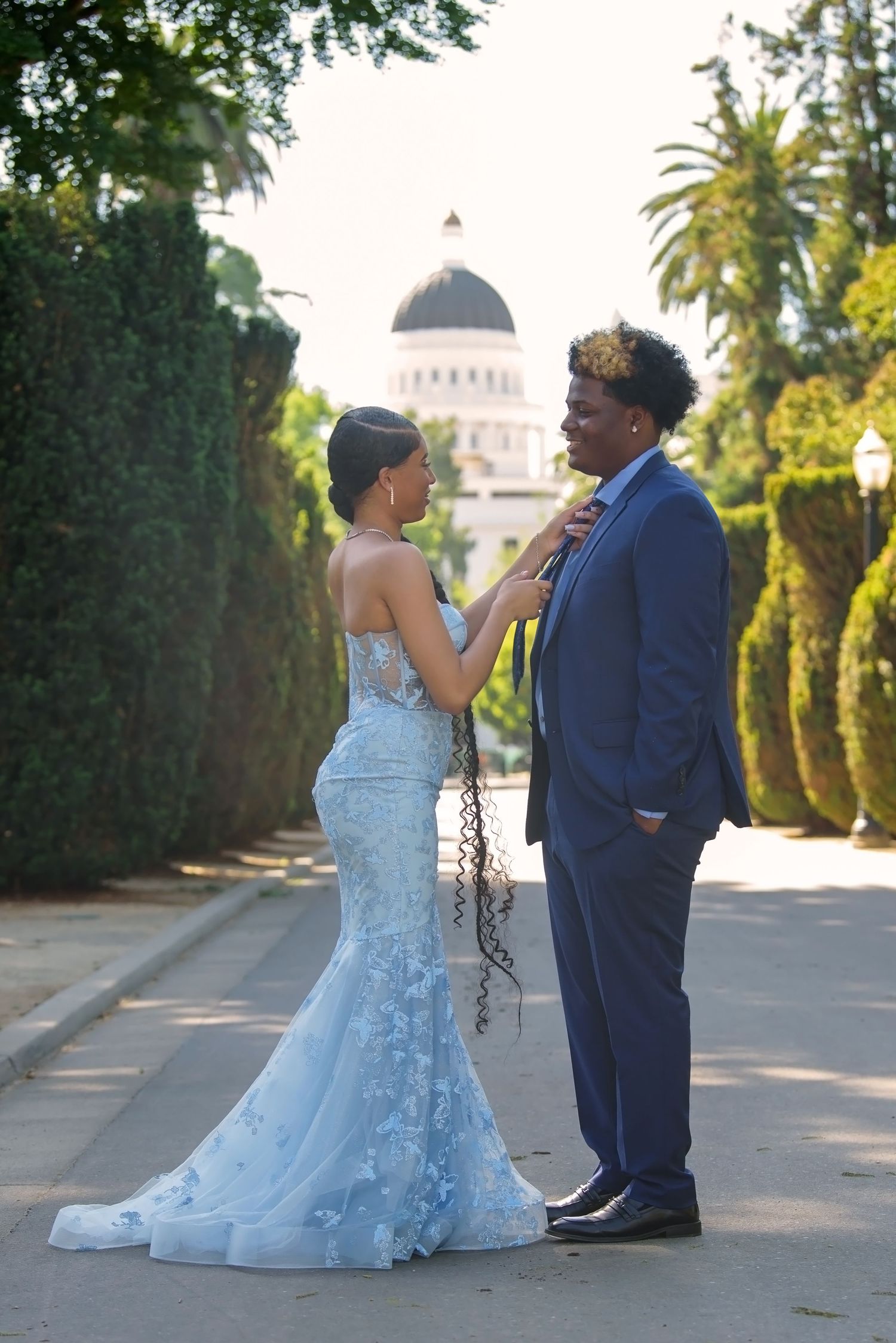 A couple in wedding attire pose together on a path with the California State Capitol dome visible in the background.