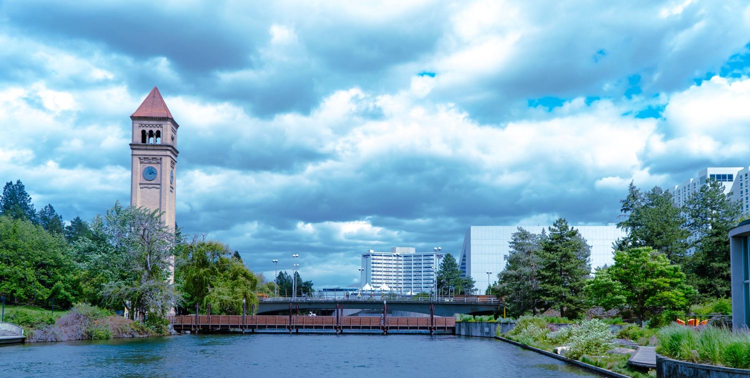 Scenic cityscape of Spokane with clock tower and bridge over river under dramatic cloudy sky.