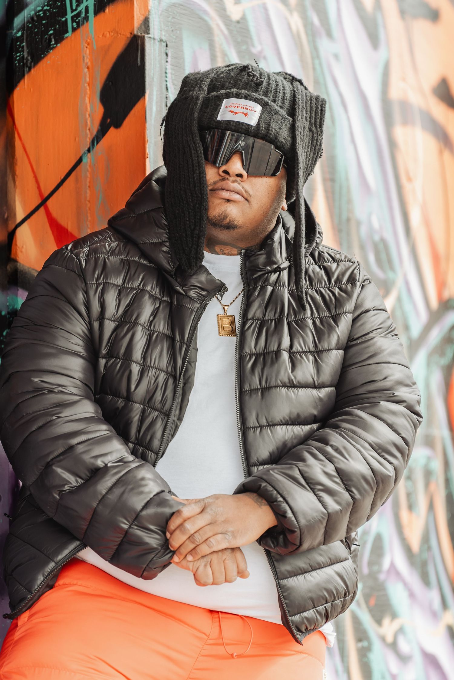 Person in a puffy grey jacket and sunglasses posing against a colorful graffiti wall background.