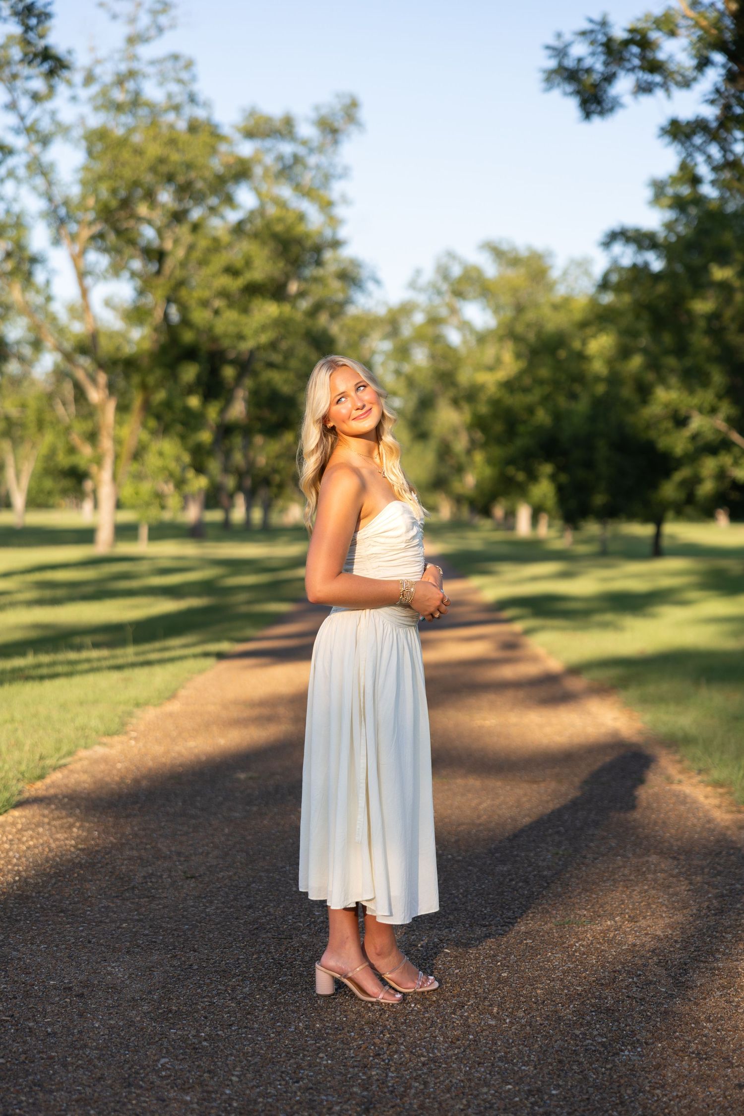 Blonde woman in white strapless dress and heels standing on a park path surrounded by green trees in golden hour light.