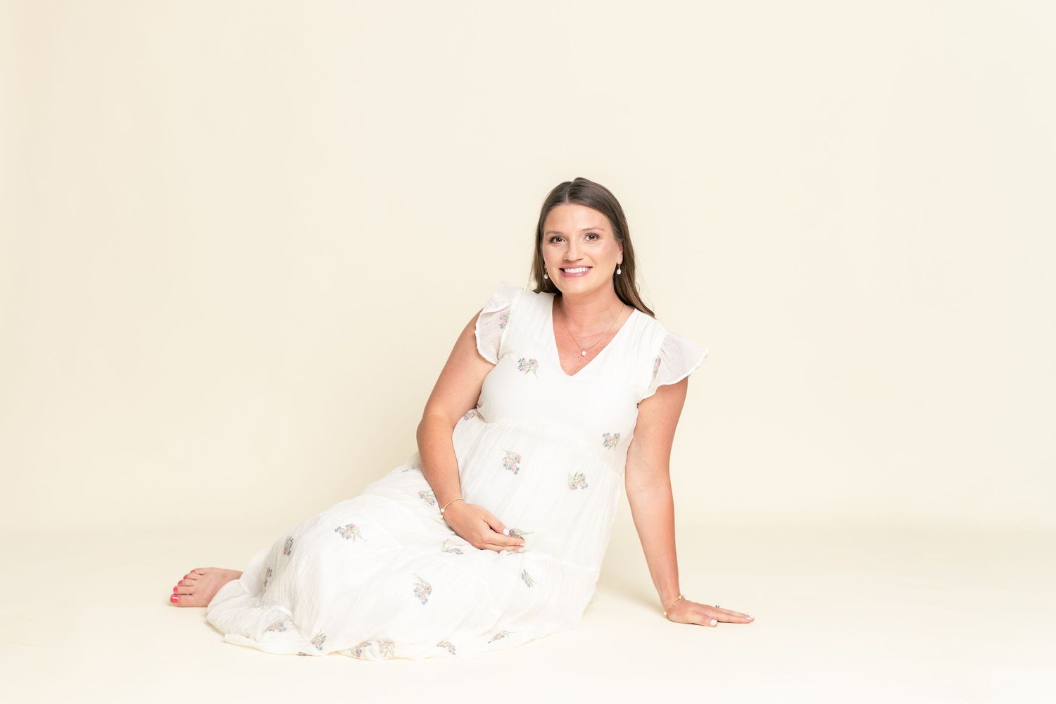 Pregnant woman in white floral dress sitting on floor, smiling softly against cream background.
