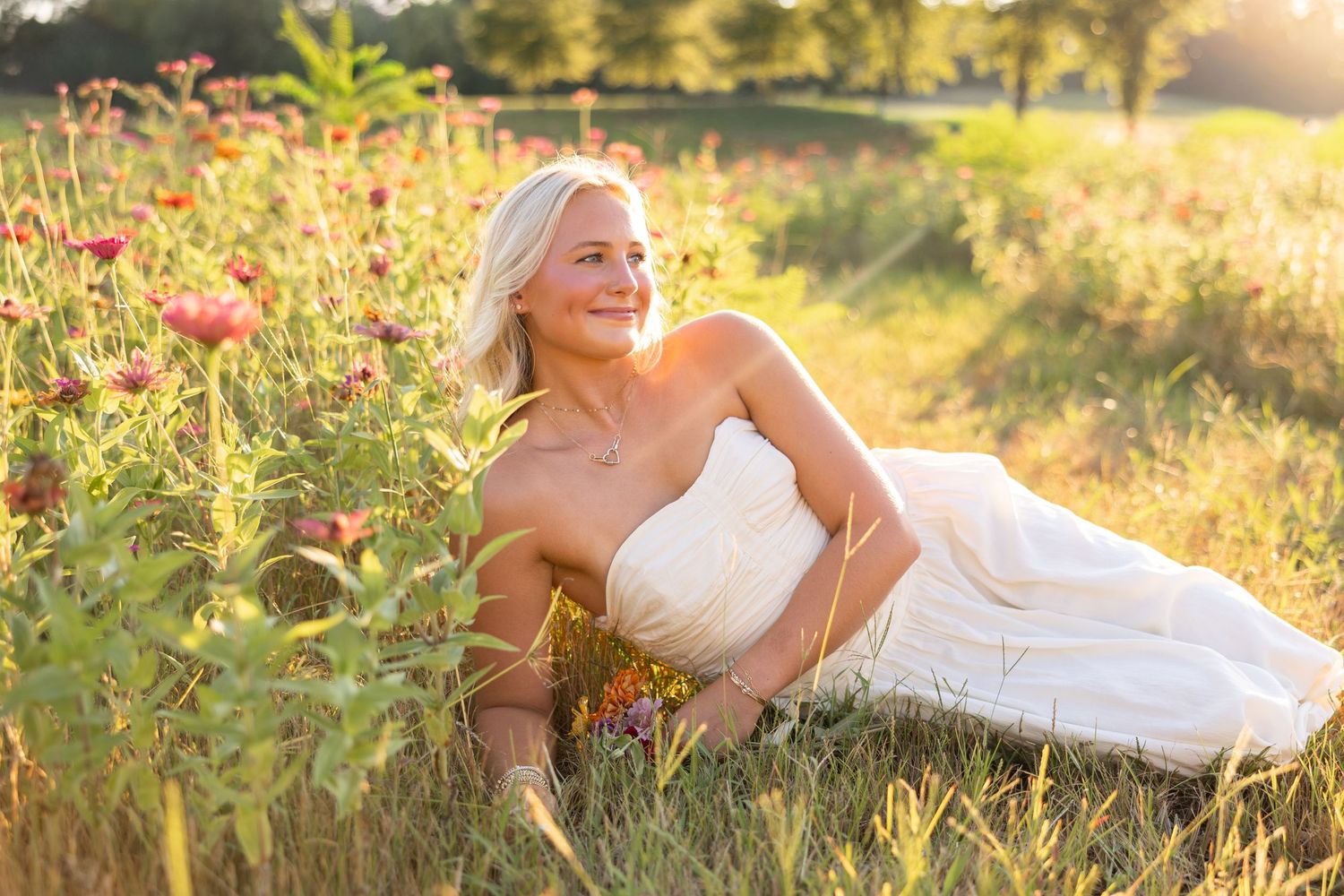 Blonde woman in white strapless gown sitting in a sunlit wildflower field, smiling peacefully in golden hour light.