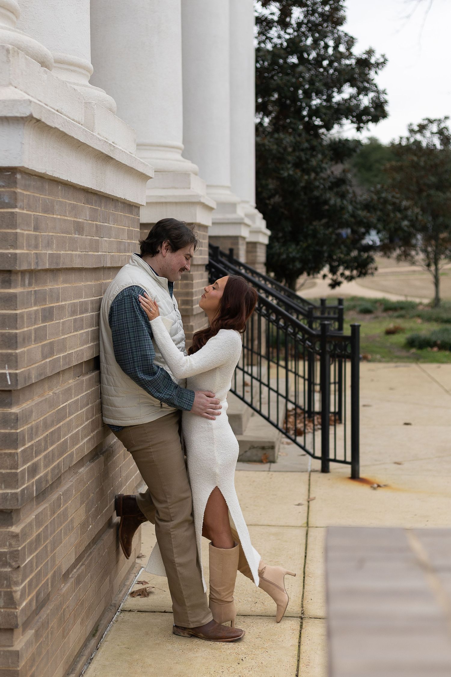 Couple embracing romantically against a brick building column, woman in white dress and boots, man in vest and khakis.
