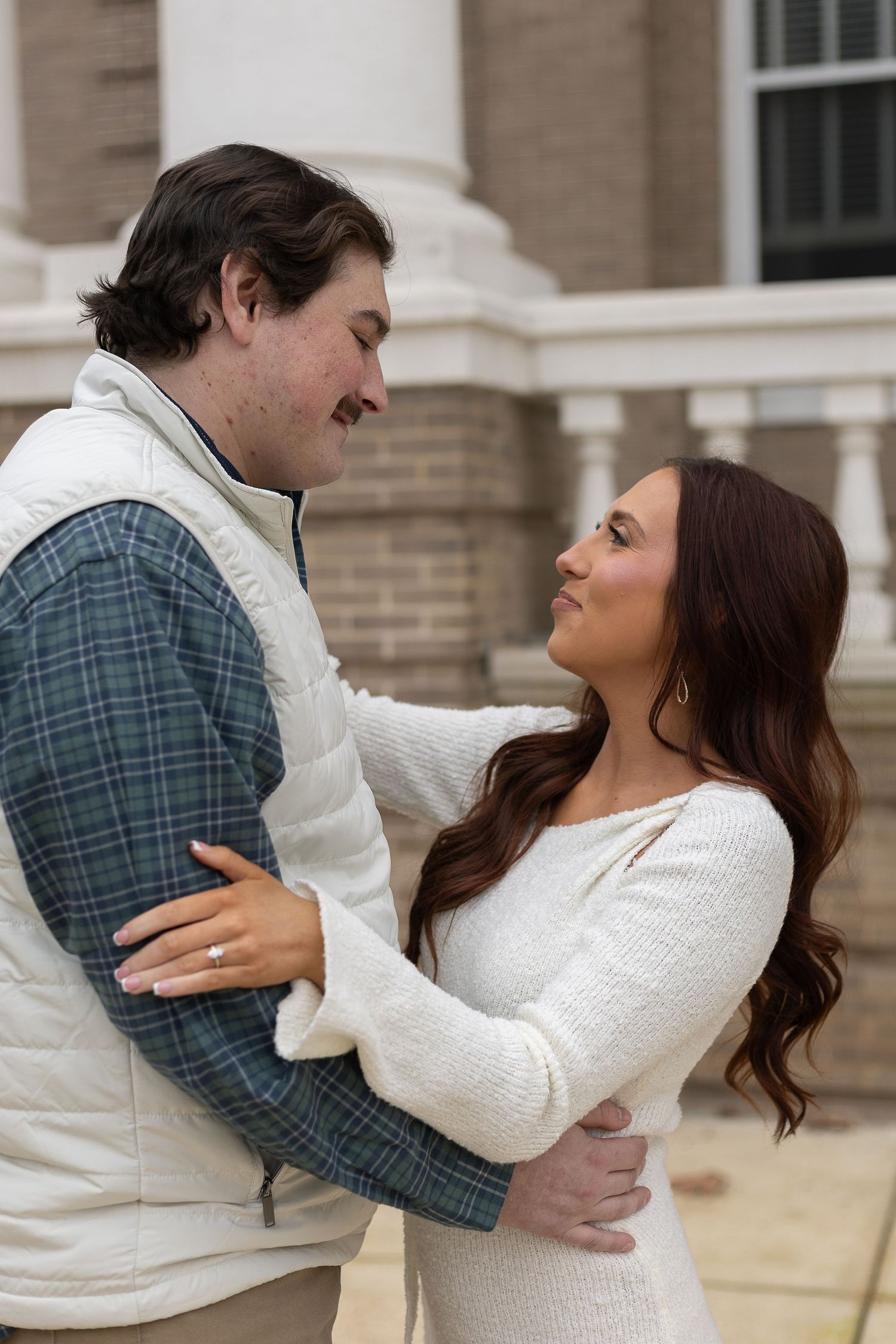 Couple embracing outdoors, woman in white sweater dress gazing up at man in plaid shirt and white vest near brick building.