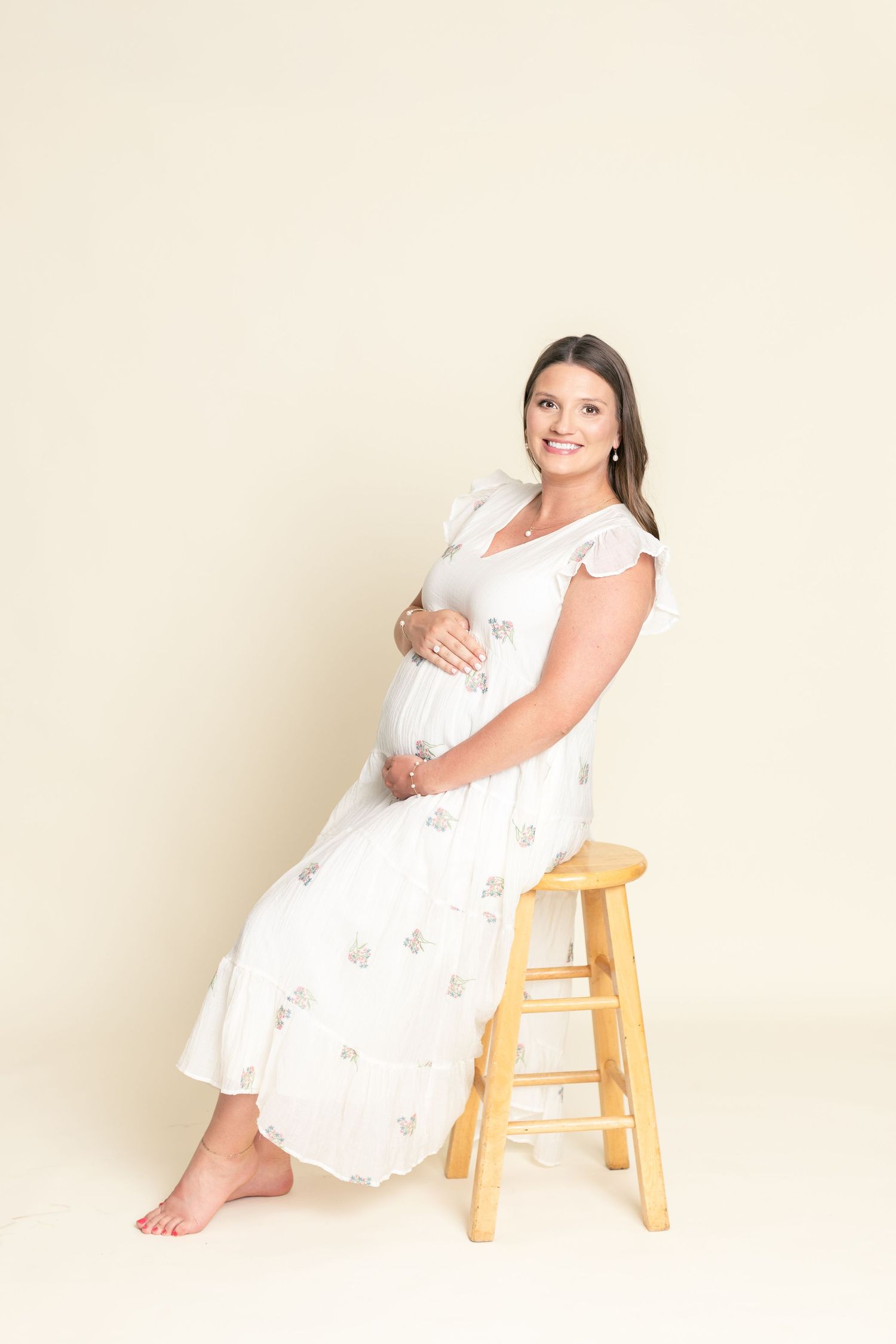 Pregnant woman in white floral dress sitting on wooden stool, cradling baby bump, smiling softly against cream background.