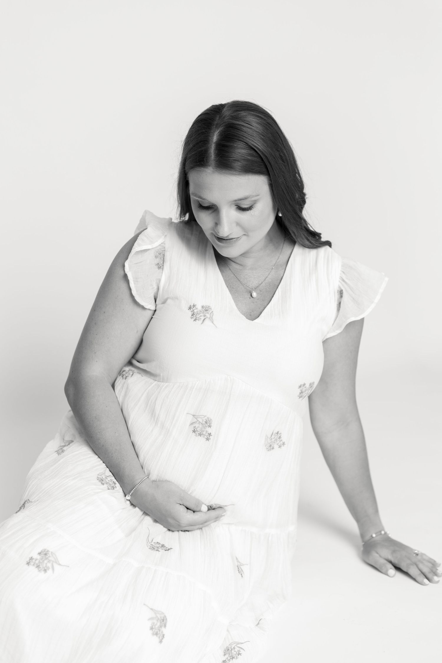 Black and white maternity photo of a pregnant woman in a white floral dress, gently cradling her baby bump.