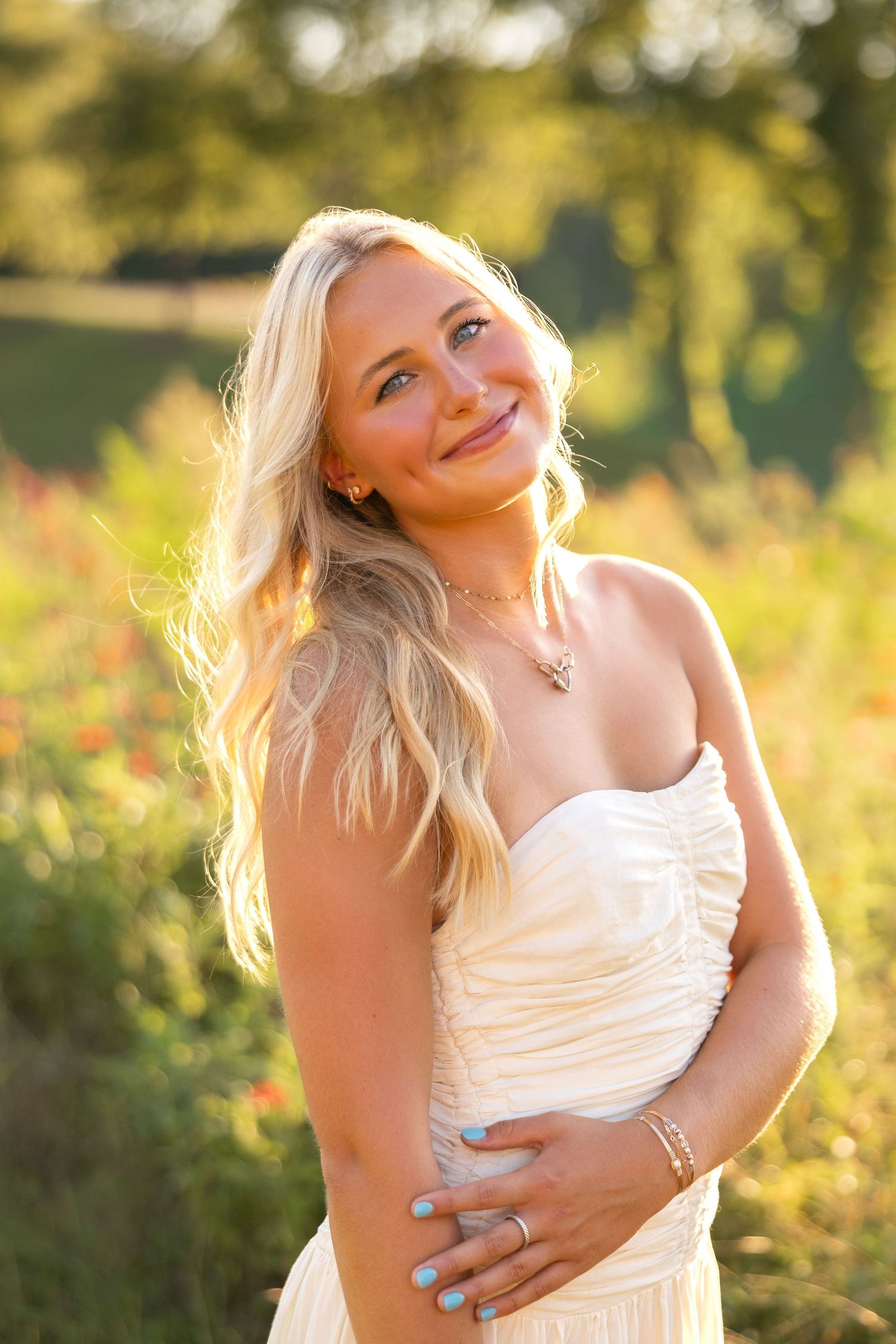 Smiling blonde woman in strapless cream dress posing outdoors in golden hour sunlight with green trees behind her.