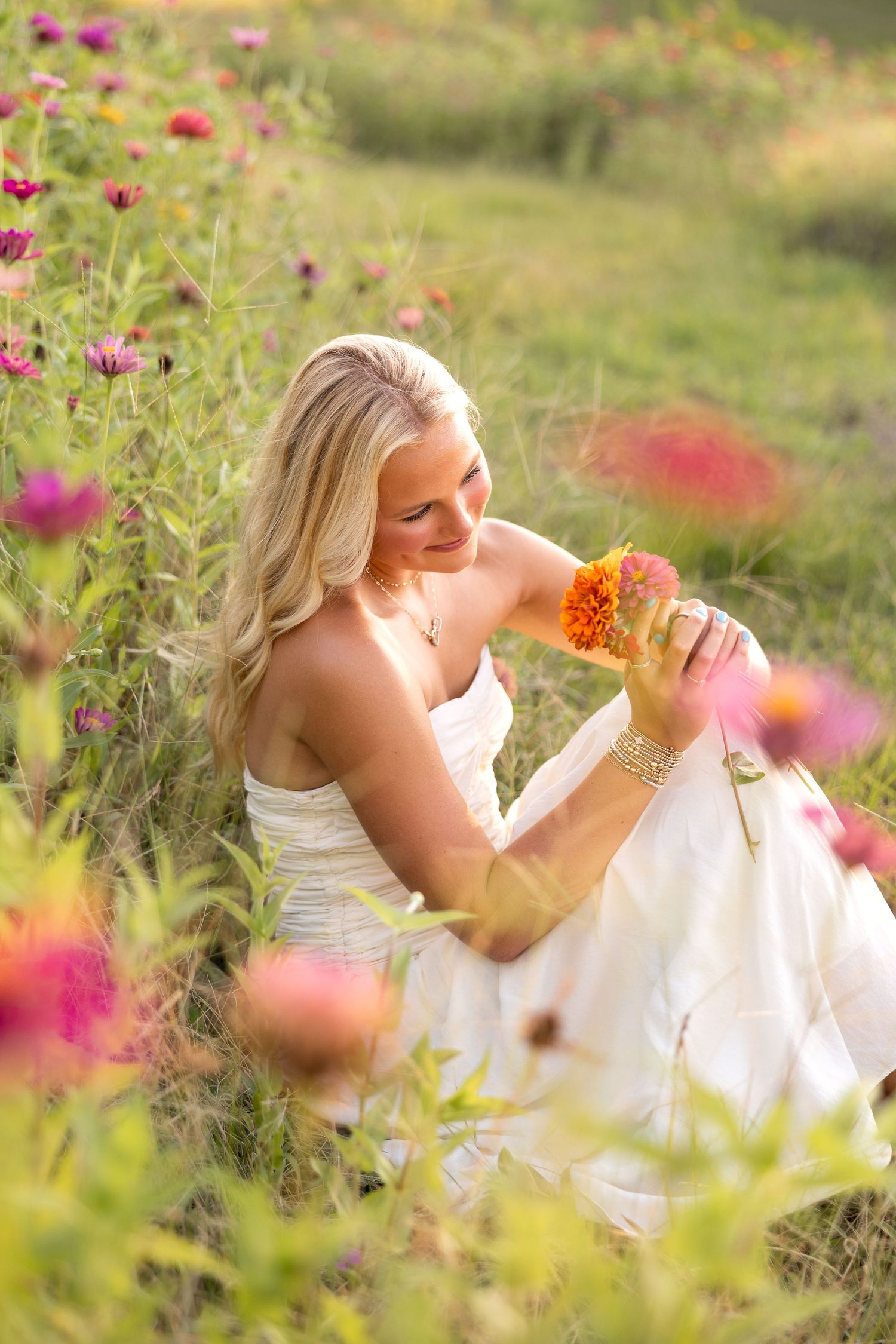 Blonde woman in white dress sitting in colorful flower field holding an orange bloom, bathed in warm golden light.