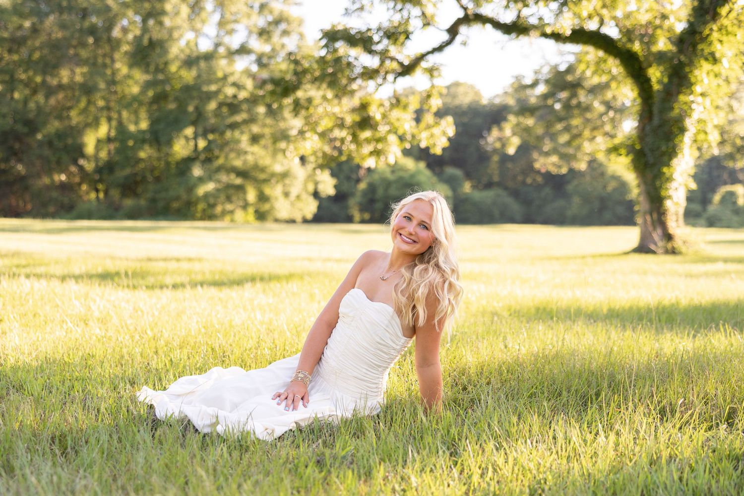 Smiling blonde woman in white dress sitting gracefully in sunlit green field with large oak tree background.