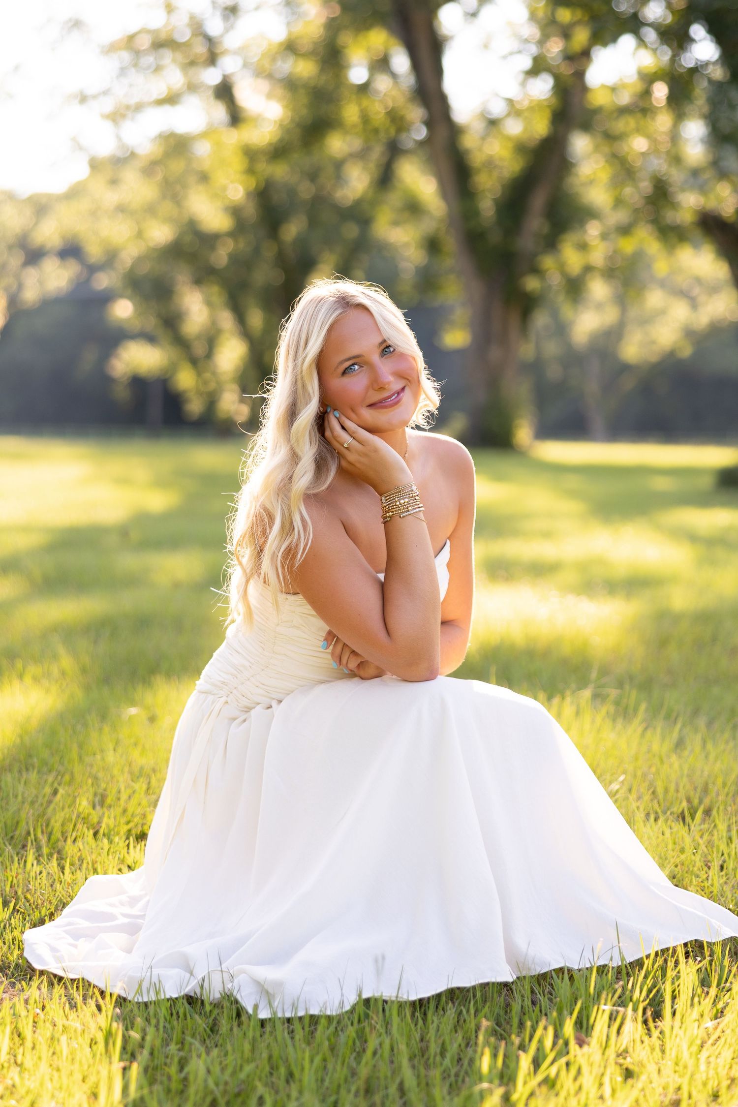 Smiling blonde bride in white gown sitting on grass in sunlit park, wearing gold bracelets.